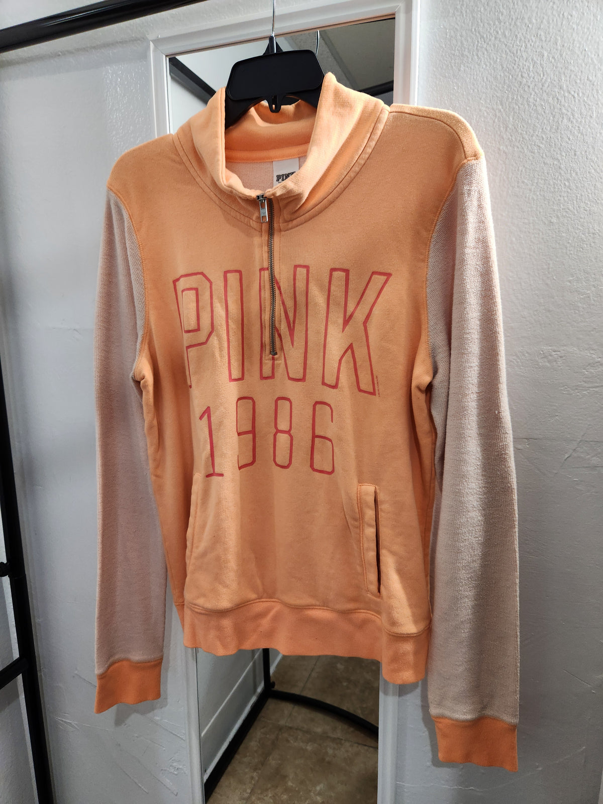 A peach and beige colorblock hoodie with 'PINK 1986' printed in red outline text on the front, hanging on a black hanger against a white door.