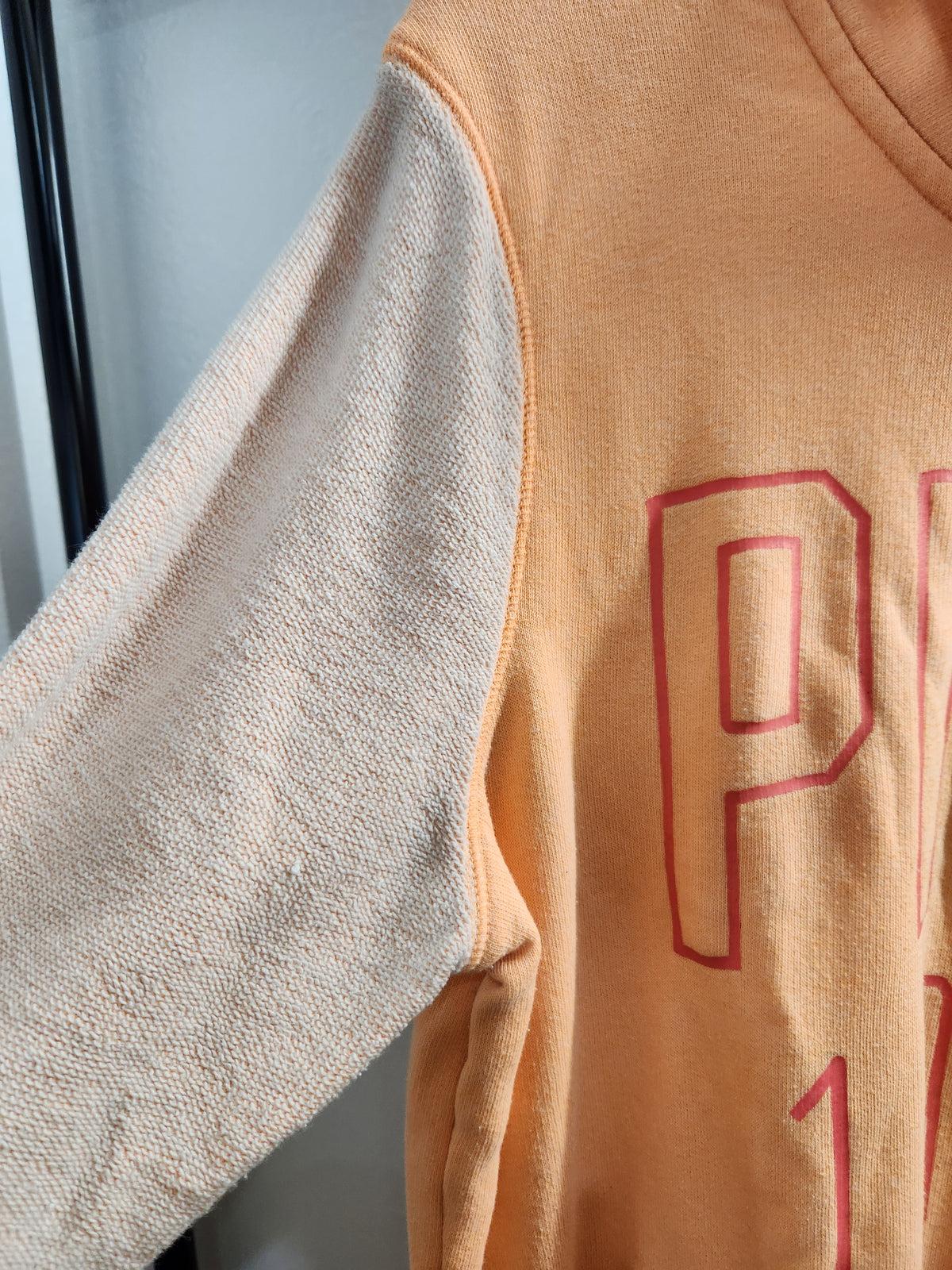 A peach and beige colorblock hoodie with 'PINK 1986' printed in red outline text on the front, hanging on a black hanger against a white door.