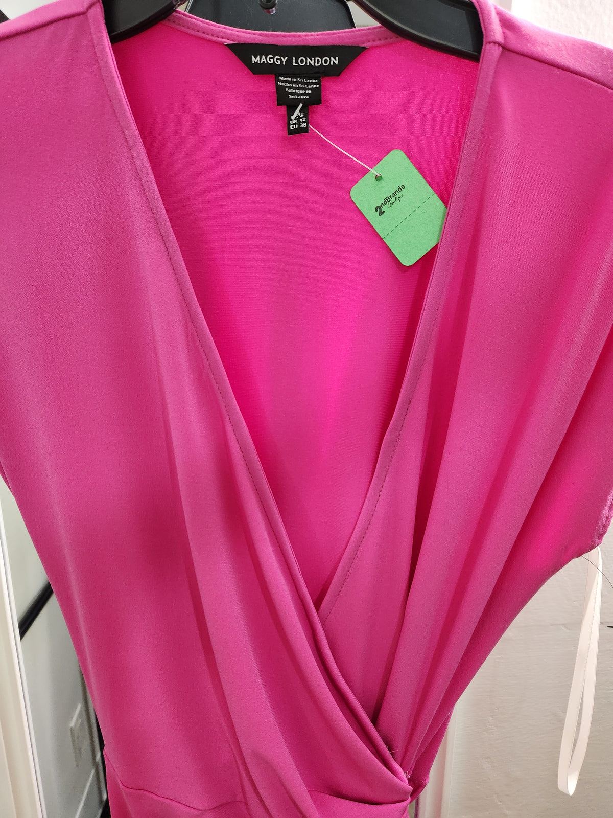 A bright pink wrap-style top or dress by Maggy London, hanging on a black hanger against a light indoor background. A black neck label and a green hangtag are visible at the neckline.