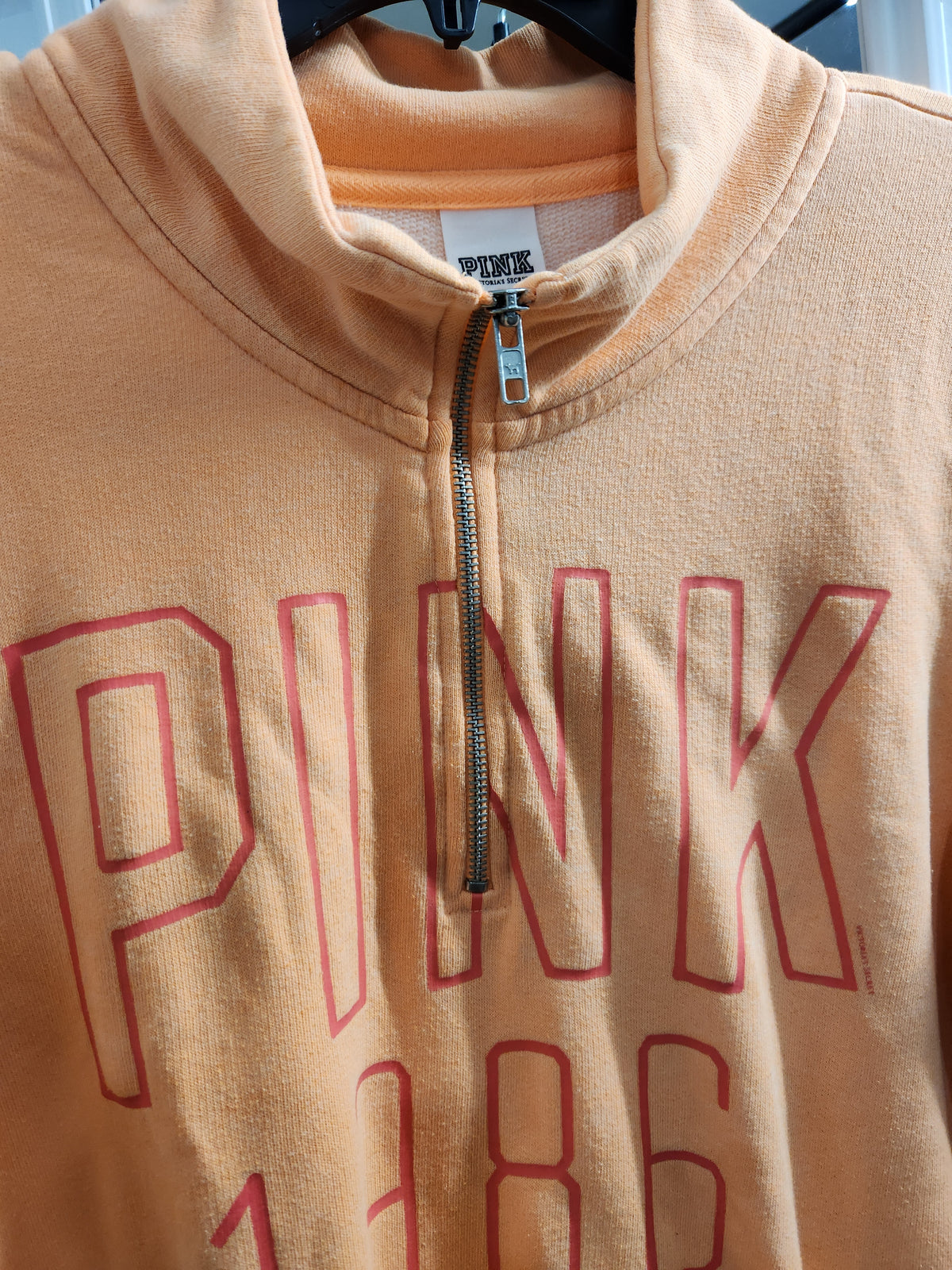 A close-up of a peach-colored PINK brand half-zip hoodie with a red outline 'PINK' graphic and '1986' printed on the front, hanging on a black hanger.