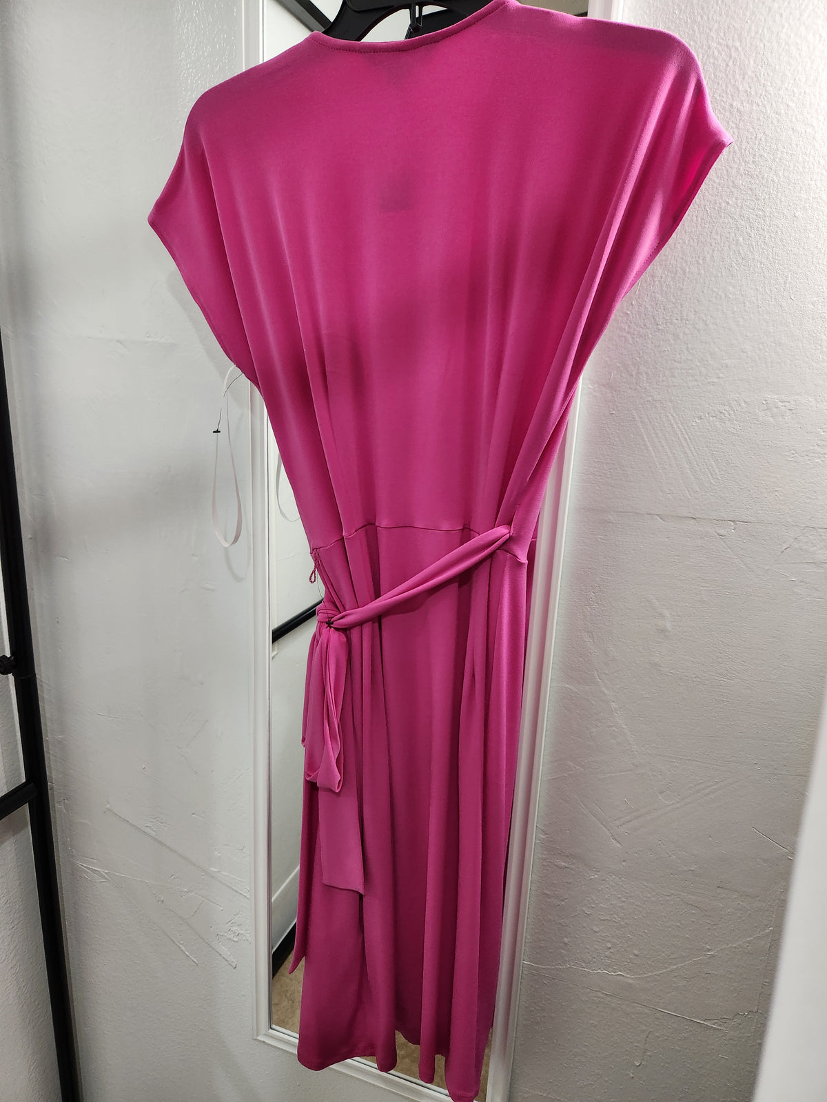 A bright pink short-sleeve wrap dress with a tie waist, hanging on a black hanger against a white wall.