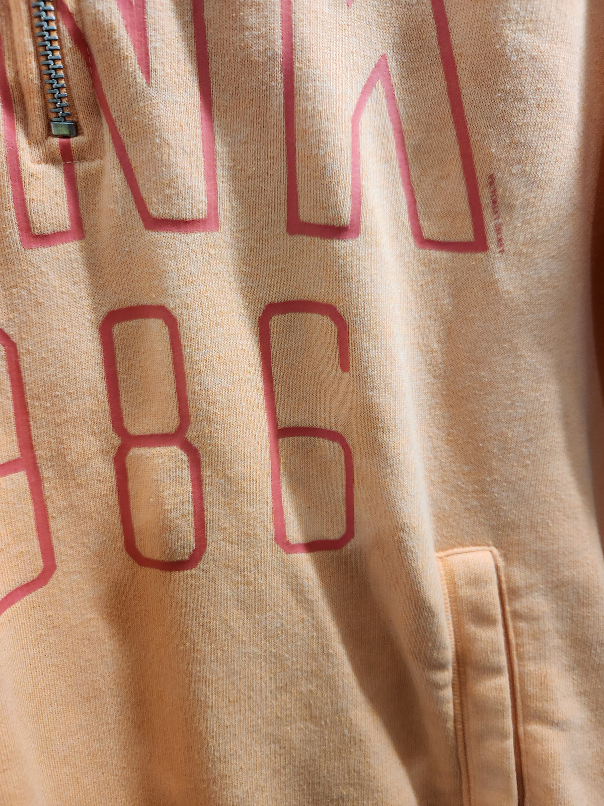 Close-up of a beige or peach-colored sweatshirt with a pink outline graphic print showing the letters 'N' and '86' and a zipper at the top left.