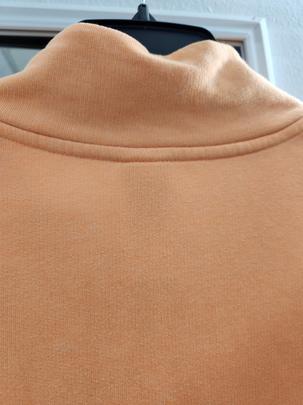 Close-up of a peach-colored knit turtleneck sweater hanging on a black hanger against a white wall and window frame.