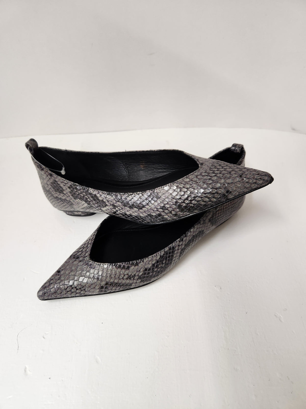A pair of pointed-toe flats with a metallic silver and black snakeskin pattern, photographed on a white surface against a white background.