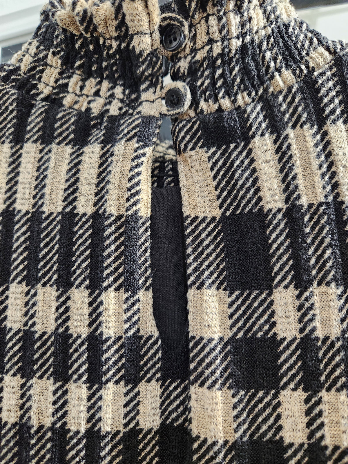 Close-up of a black and beige plaid knit garment with a high gathered neckline and dark buttons.