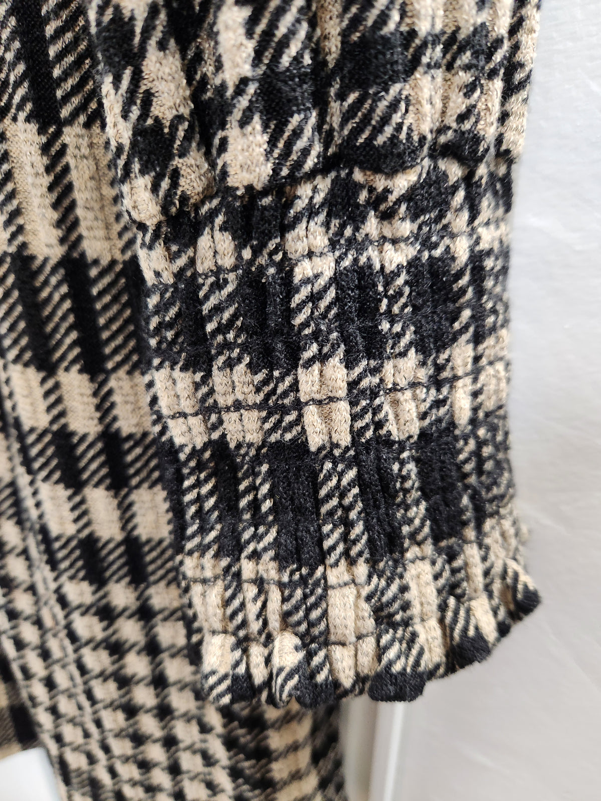 Close-up of a black and beige plaid tweed fabric with pleated detailing along one edge, shown against a light neutral background.