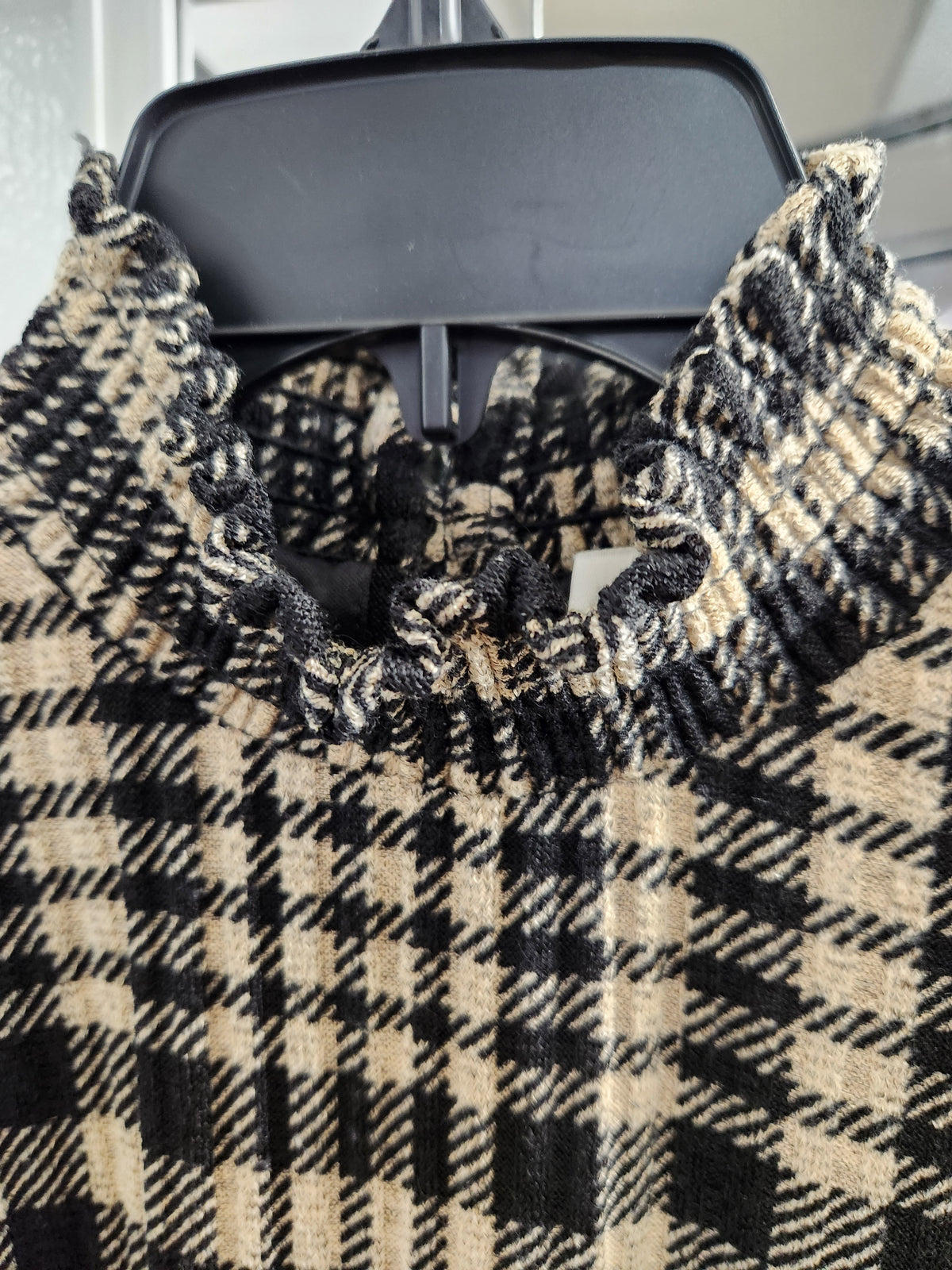 A close-up of a black and beige plaid knit top with a gathered ruffled mock neck, hanging on a black plastic hanger.