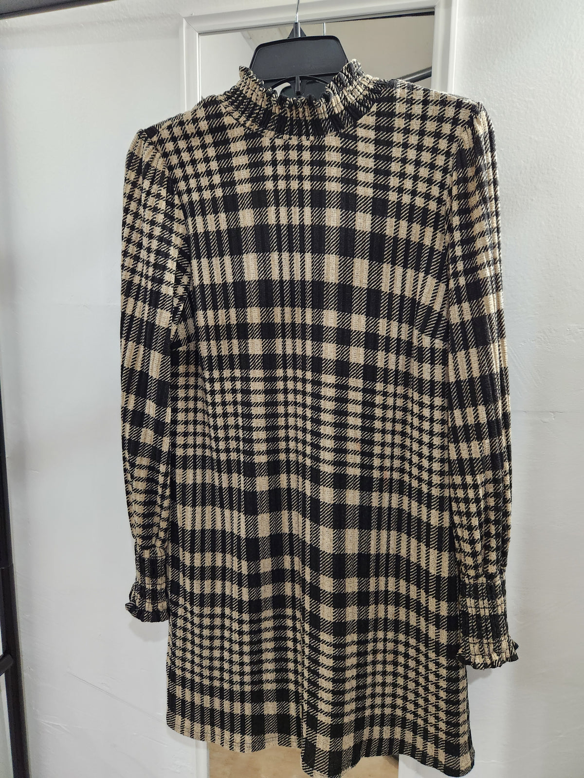 A long-sleeve mock neck tunic top with a black and beige plaid pattern, hanging on a black hanger against a white door.