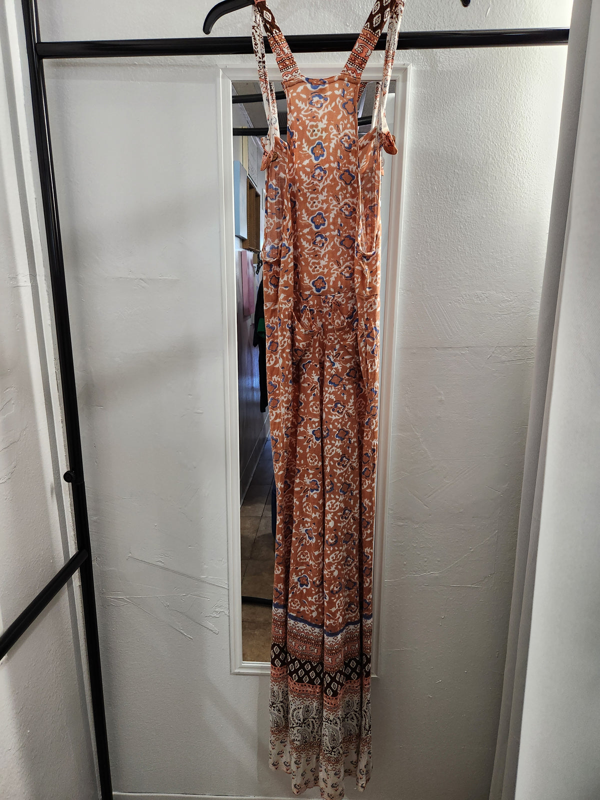A sleeveless bohemian-style maxi dress with a rust-orange, blue, and white pattern, hanging on a black clothing rack in front of a white wall.