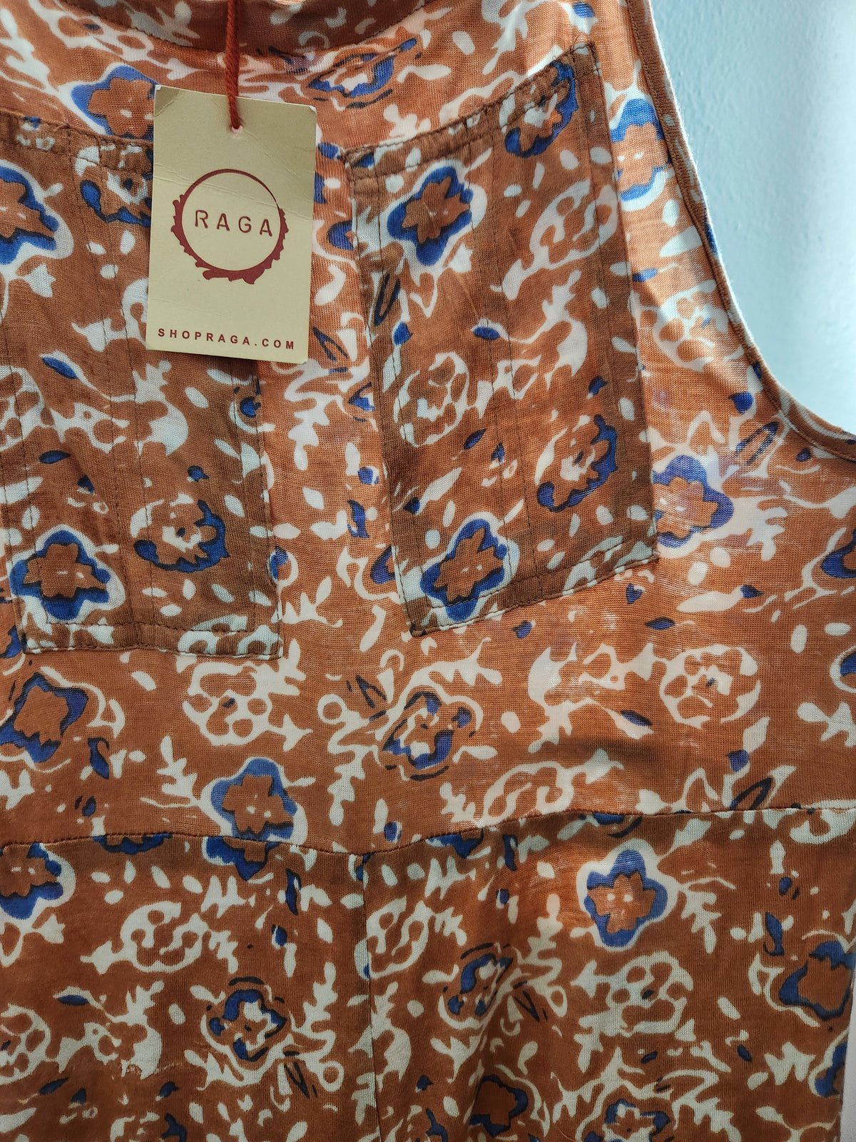 Close-up of a sleeveless top or dress with an abstract floral print in rust orange, cream, and blue. A hanging paper tag reads RAGA SHOPRAGA.