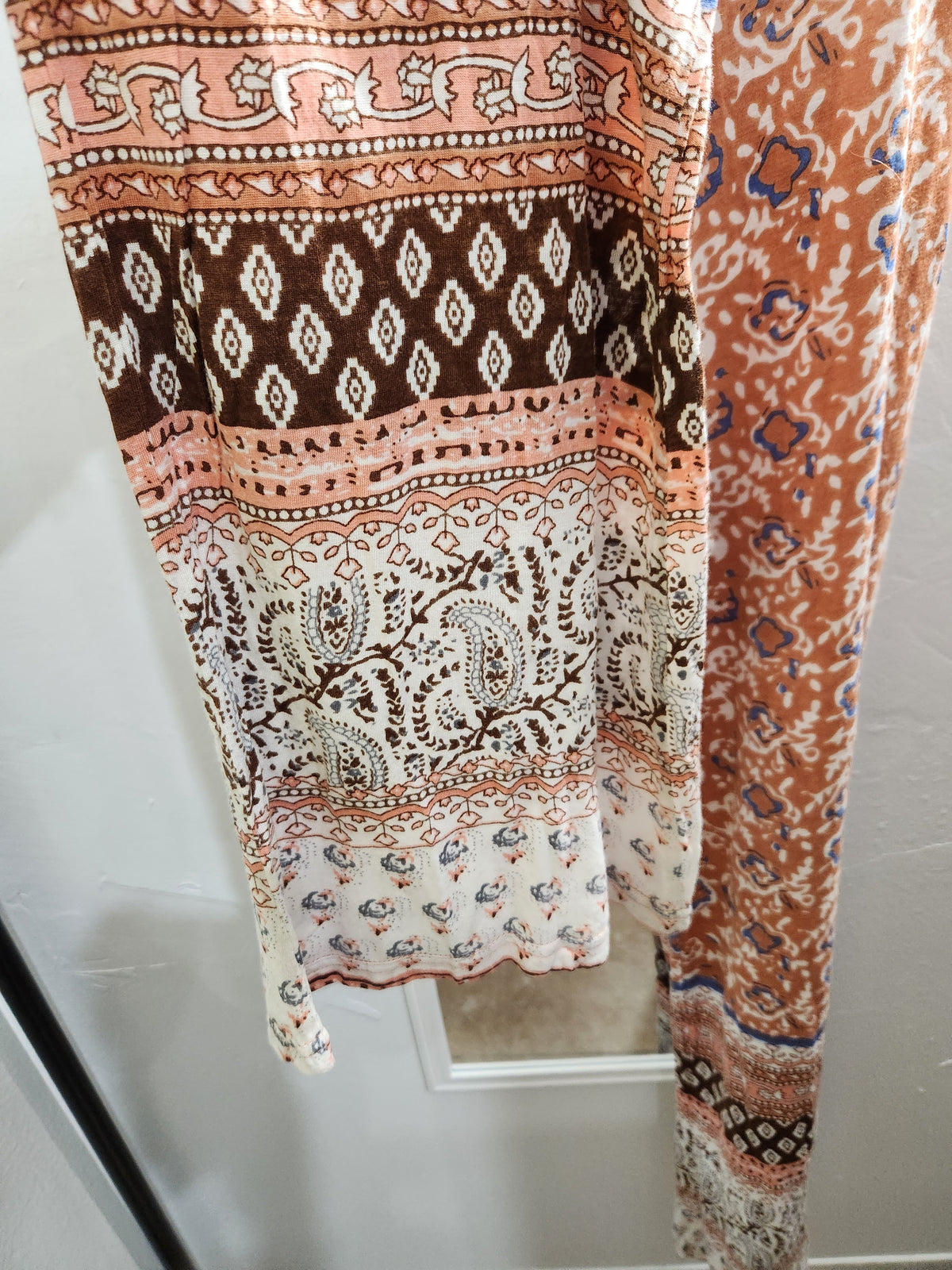 A close-up of a patterned fabric garment, likely a scarf, shawl, or section of a dress, featuring multiple horizontal bands of paisley, diamond, and floral motifs in peach, brown, cream, and blue.
