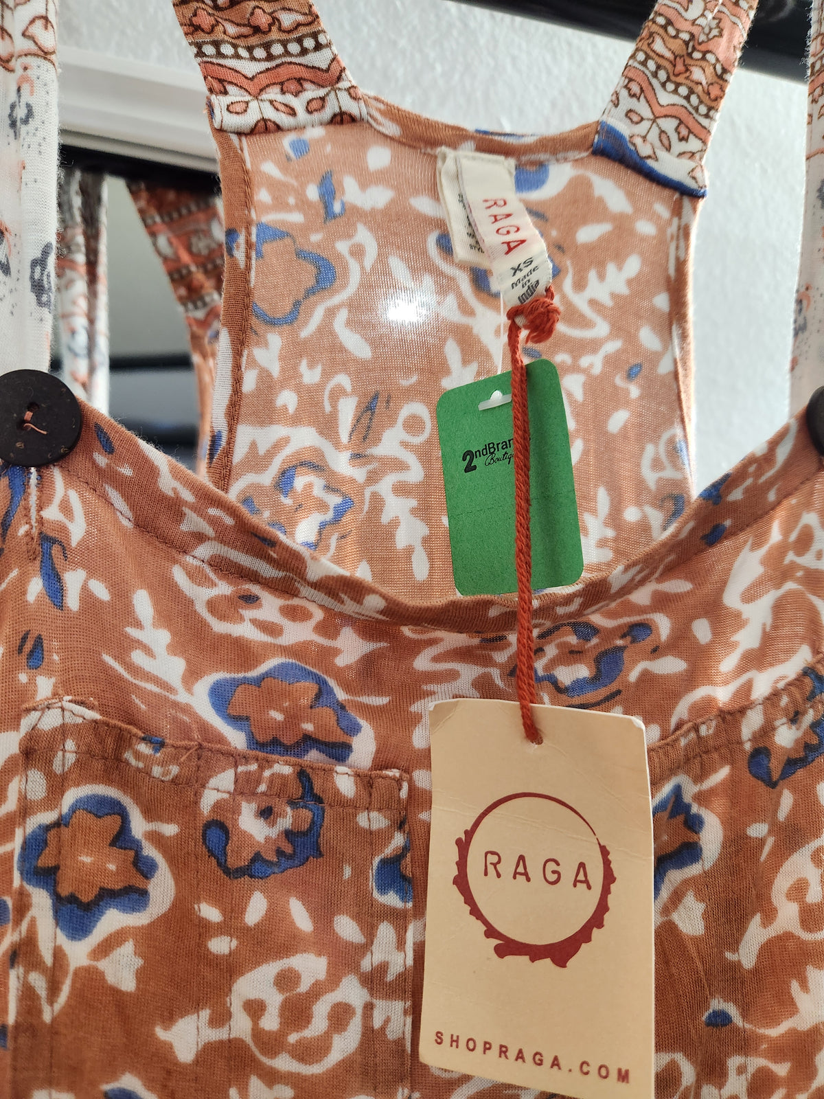 Close-up of a RAGA brand patterned garment, likely a dress or romper, featuring a rust-orange, white, and blue floral print. The image shows the chest area with a button, a front pocket, and a hanging paper tag and green product tag.