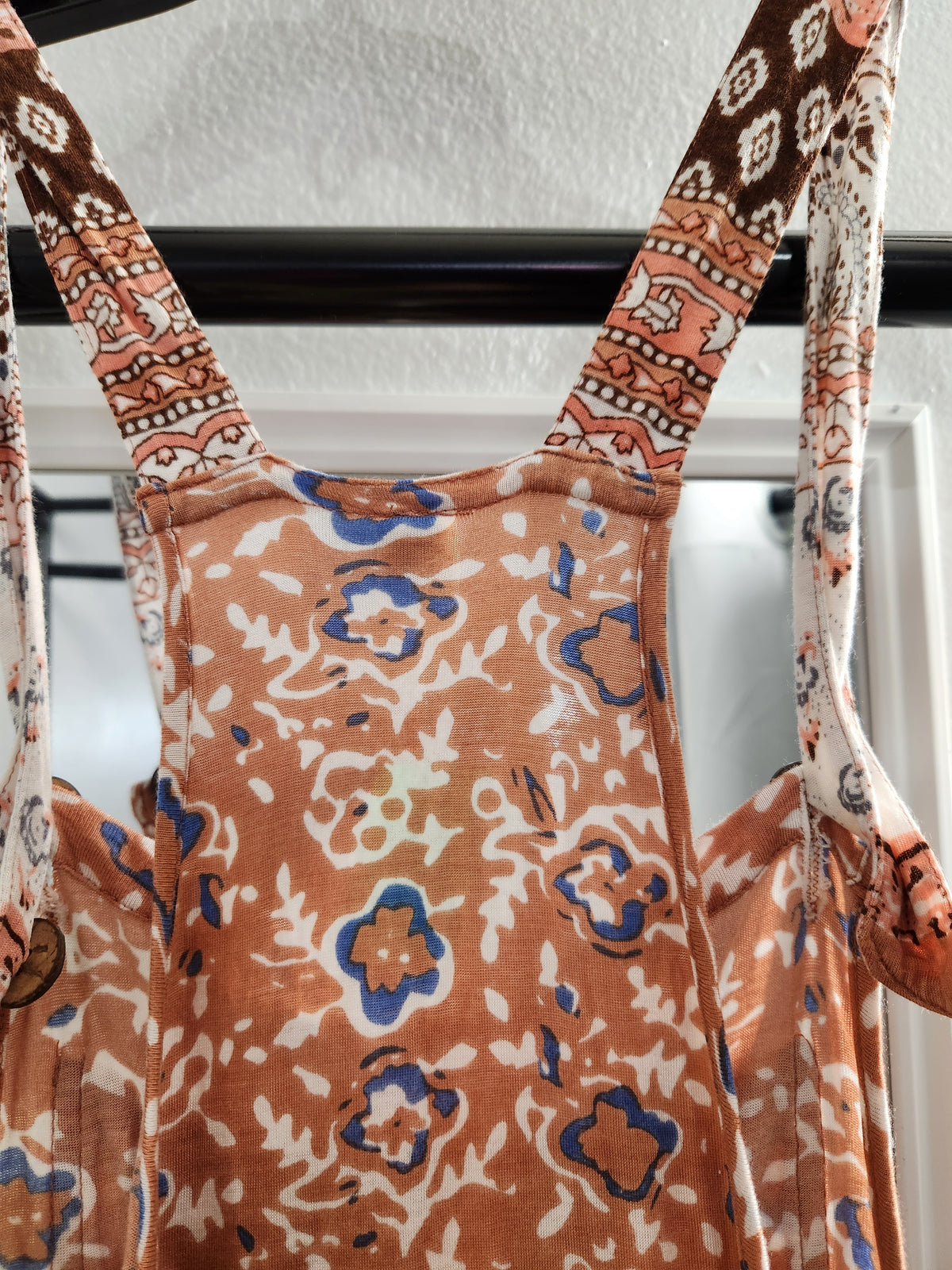 A close-up of a patterned garment or dress with wide straps, featuring an abstract floral print in terracotta orange, cream, and blue, hanging against a white wall.