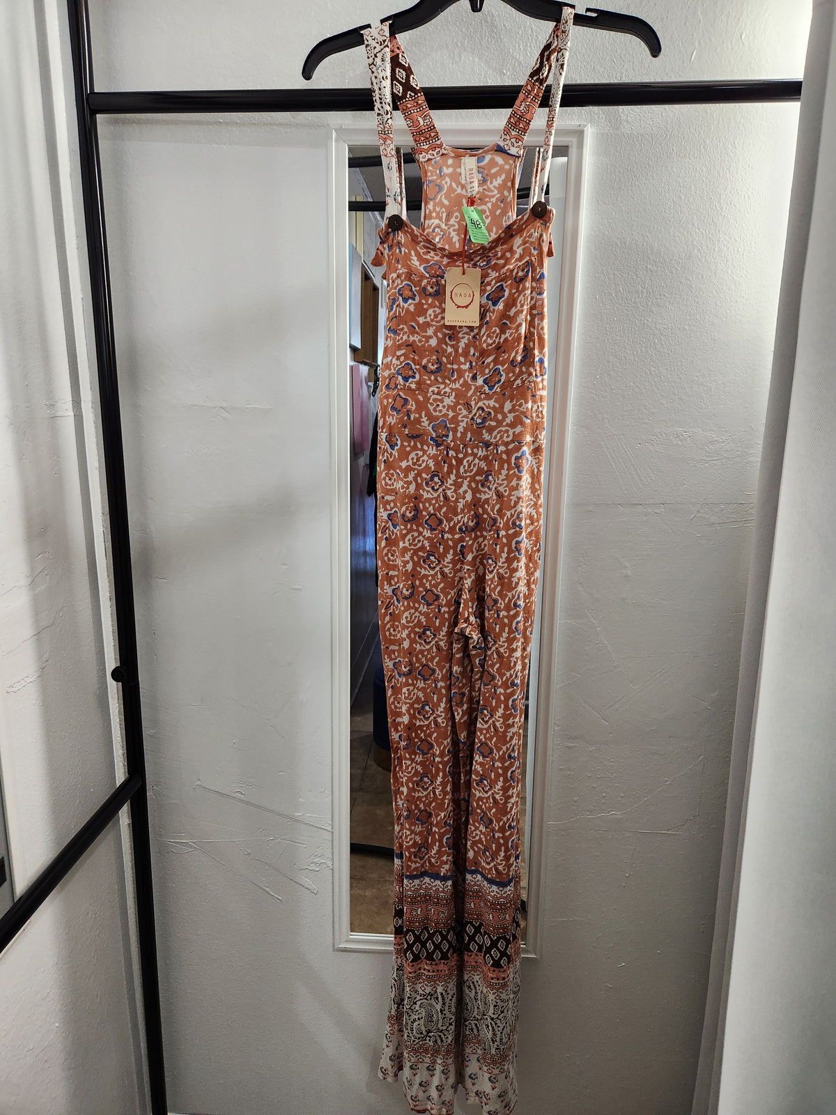 A sleeveless bohemian-style jumpsuit with an intricate orange, brown, and beige floral pattern, hanging on a black hanger in front of a white door with a mirror.