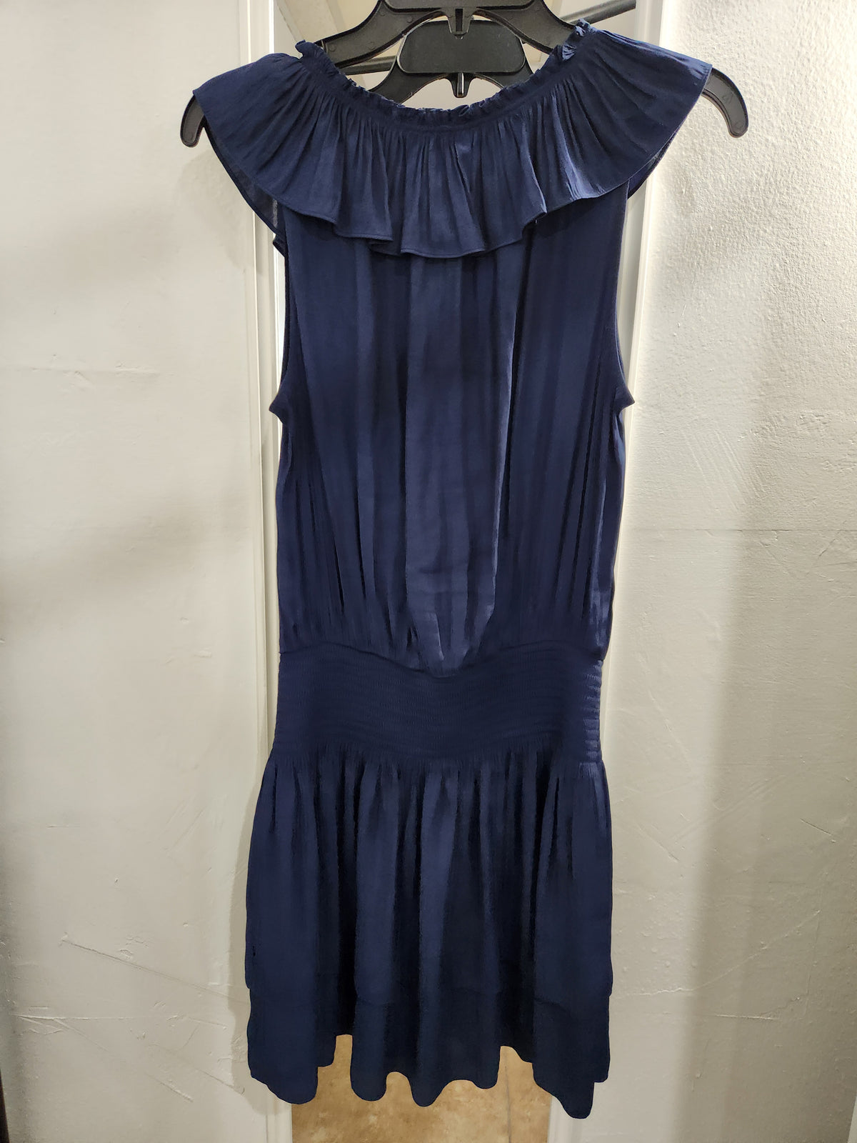 A navy blue sleeveless dress with a ruffled off-the-shoulder neckline, gathered bodice, and flared skirt, hanging on a black hanger against a light-colored wall.