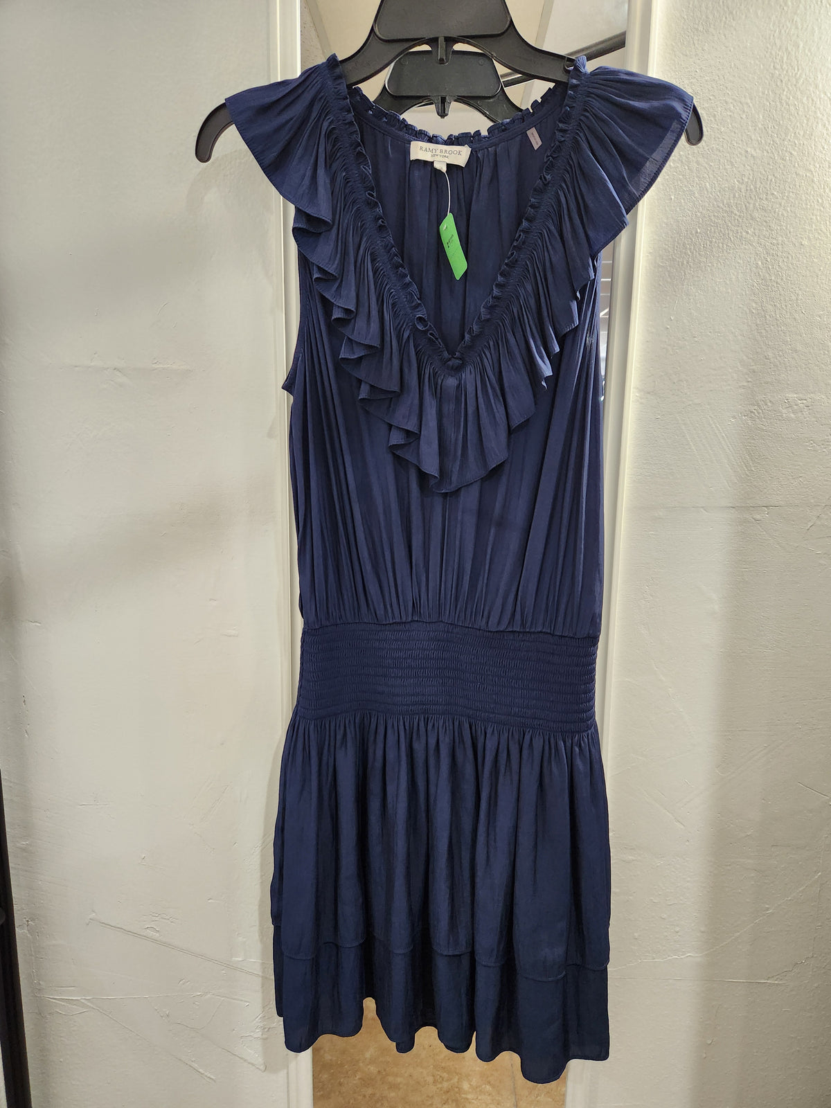 A navy blue sleeveless dress with ruffle details on the bodice and cap sleeves, hanging on a black hanger against a white paneled wall.
