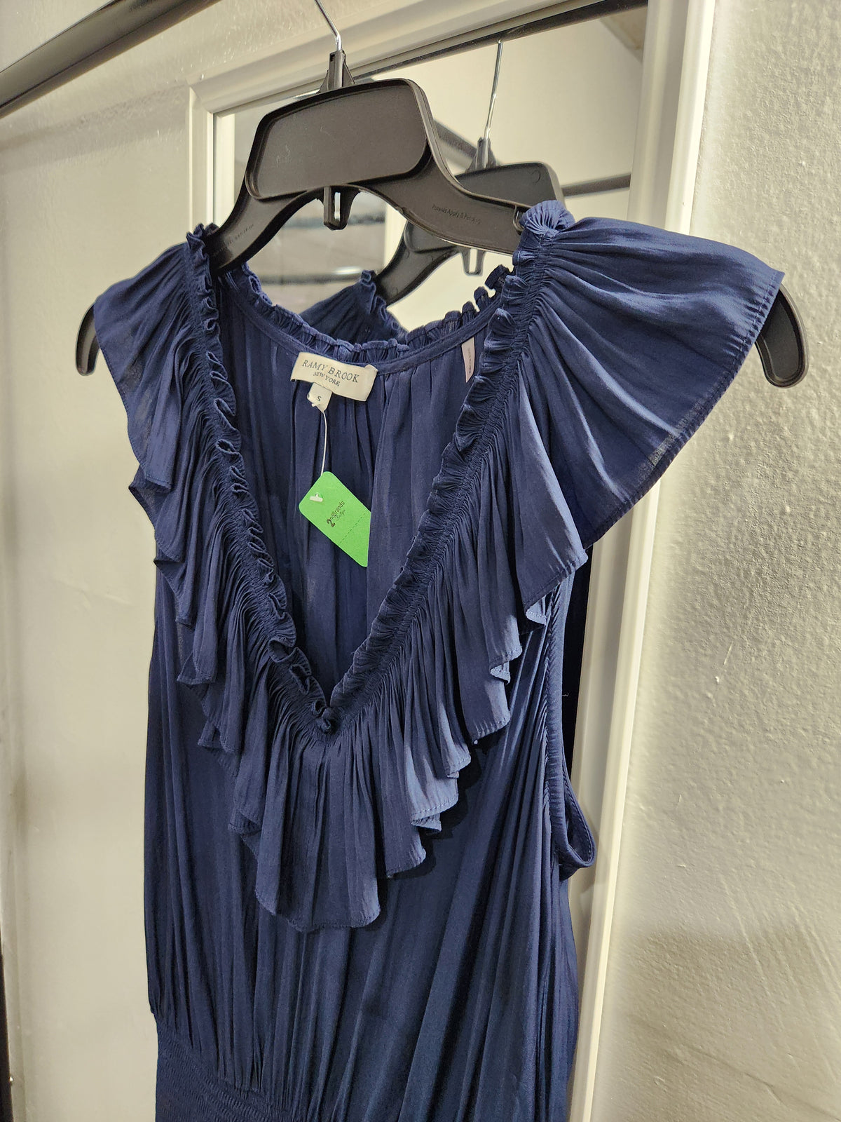 A navy blue sleeveless dress with ruffled trim and cap sleeves, hanging on a black hanger against a white door and textured wall.