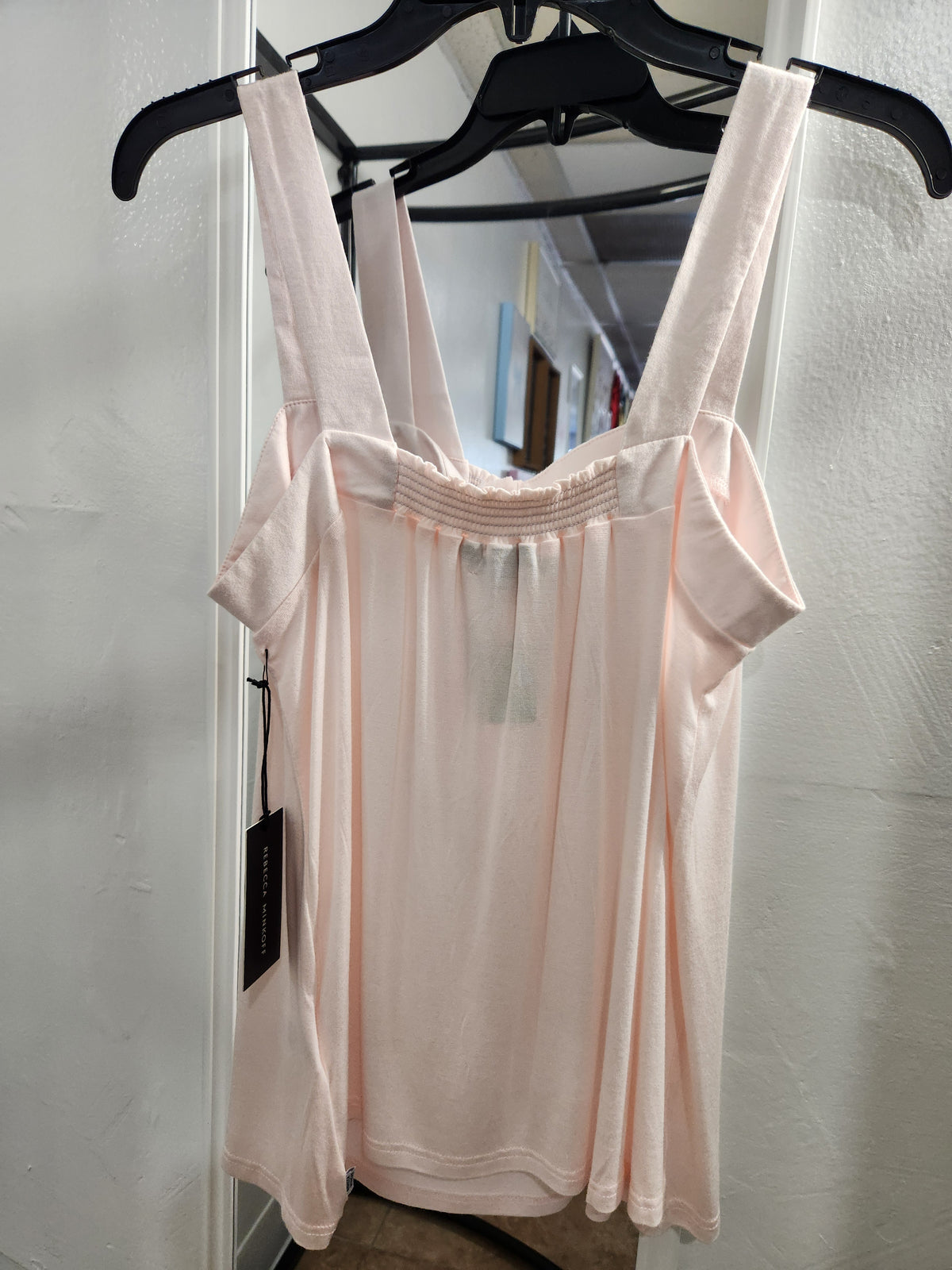 A light pink sleeveless tank top with wide shoulder straps and a smocked bodice, hanging on a black hanger against a white wall.