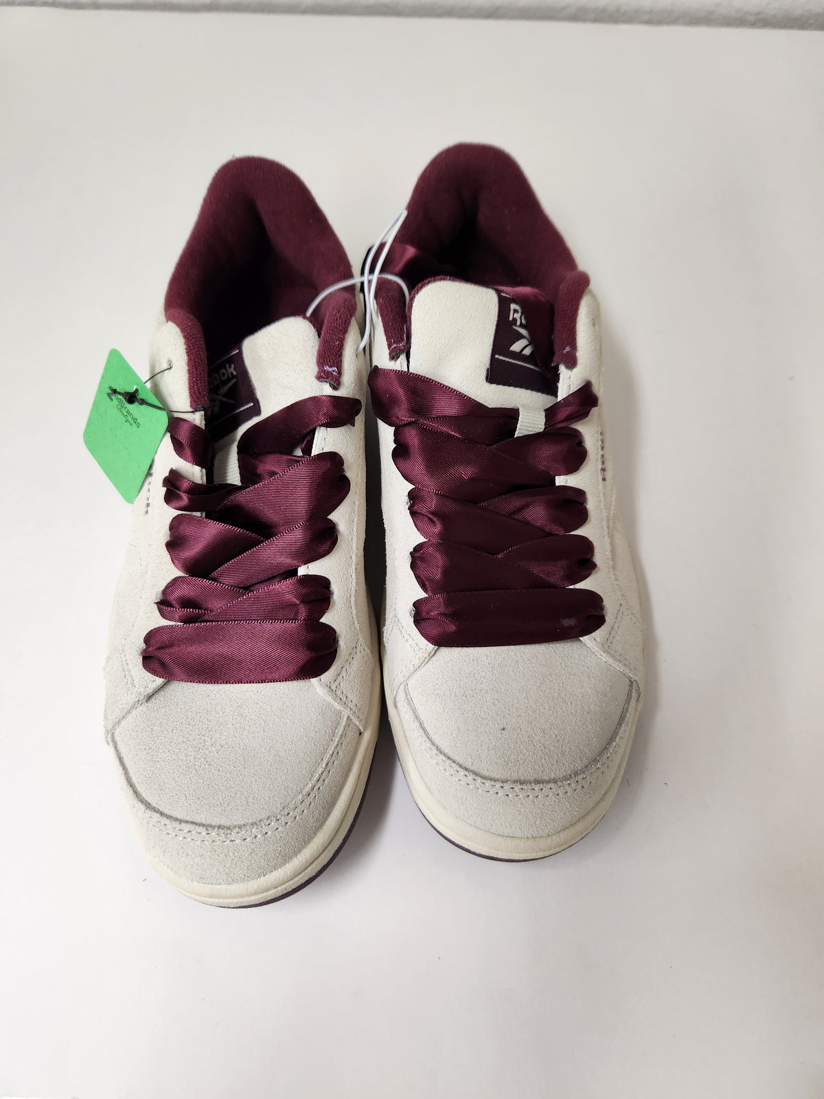 A pair of white low-top sneakers with deep burgundy satin laces and matching burgundy lining, shown on a plain white surface with a green price tag attached.