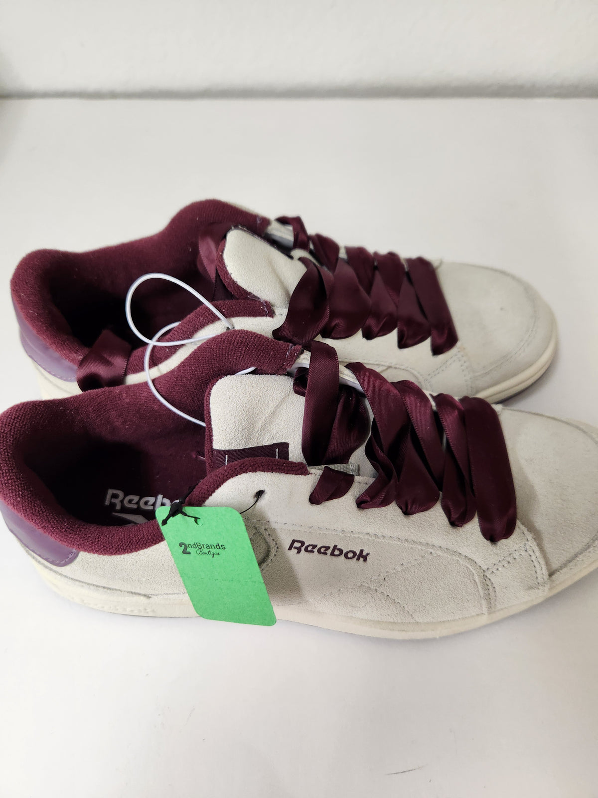 A pair of Reebok low-top sneakers in off-white suede with dark burgundy satin laces and matching burgundy inner lining, shown on a white surface with a green retail tag attached.

