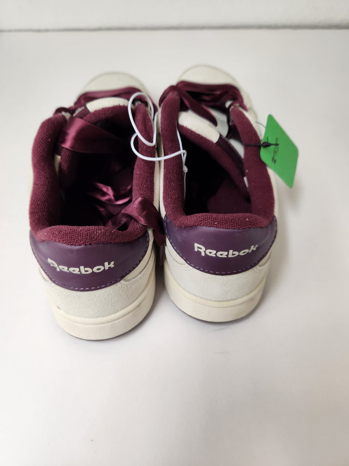 A pair of Reebok sneakers shown from the back, featuring a cream-colored suede upper, burgundy satin laces, and a burgundy heel tab with the Reebok logo. A green price tag is attached to the lace.