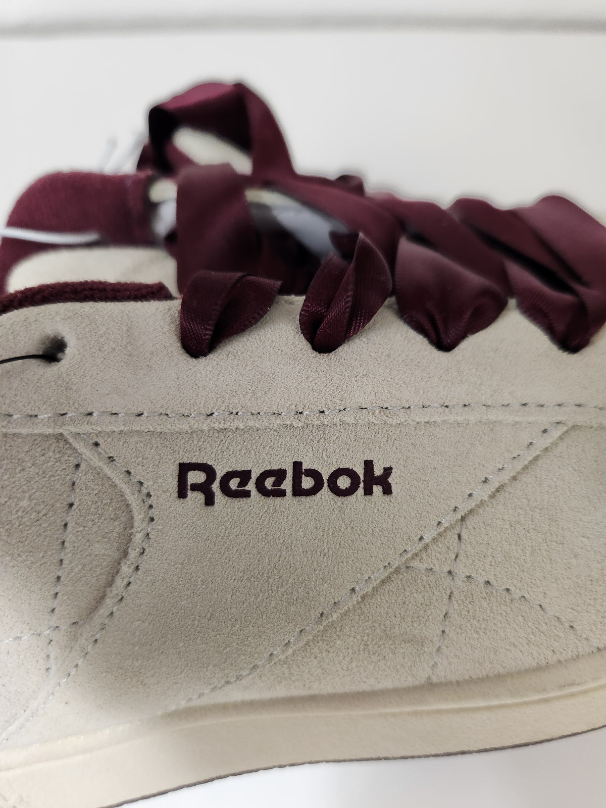Close-up of a Reebok sneaker featuring a light beige suede upper, dark burgundy ribbon laces, and the Reebok logo printed on the side.