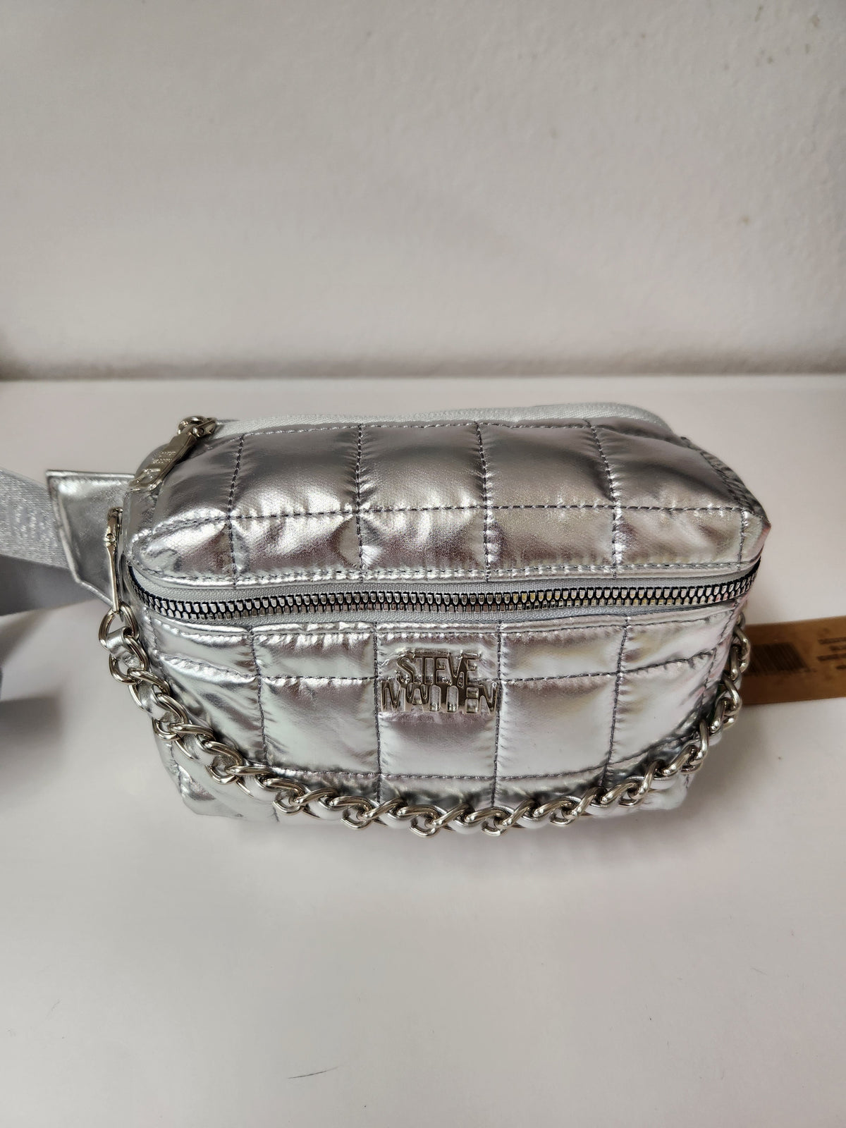 A metallic silver quilted crossbody bag with a silver chain strap and a silver-tone metal logo plate on the front, photographed on a white surface against a white wall.