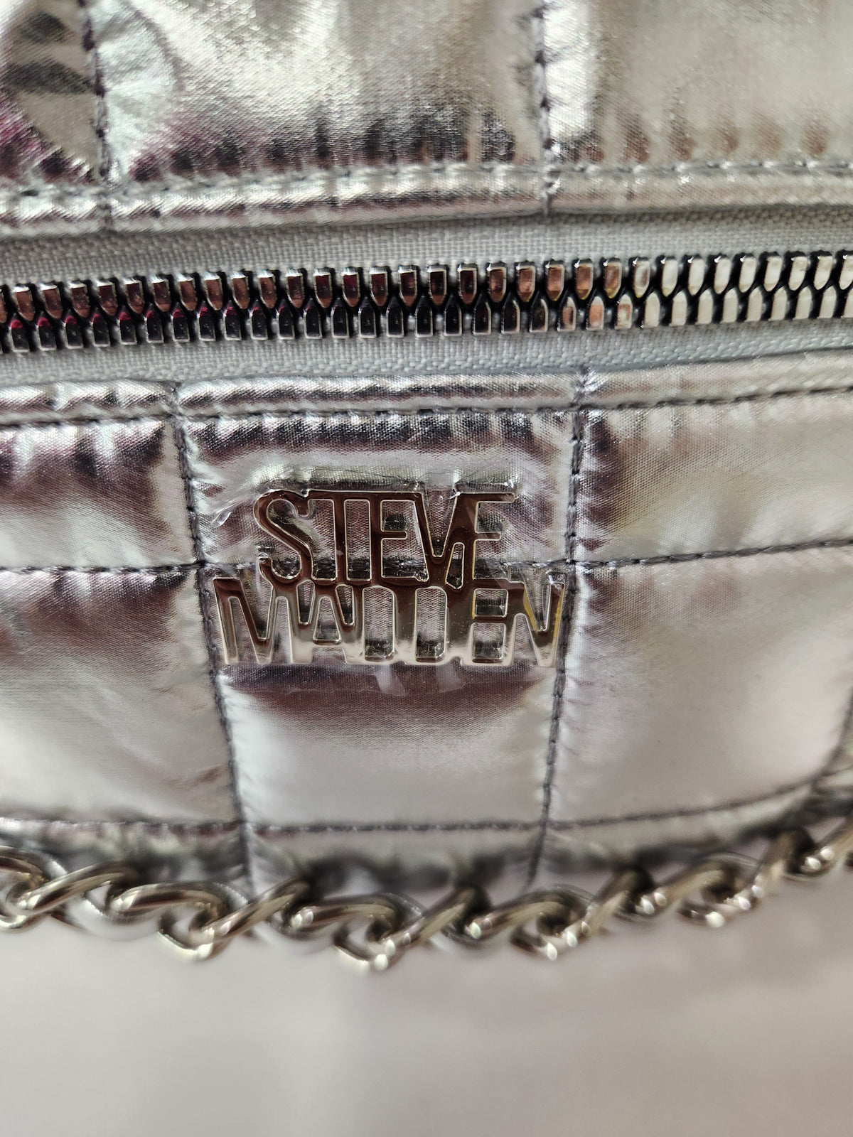 Close-up of a metallic silver quilted bag with a silver chain strap and a prominent 'STEVE MADDEN' metal logo plaque on the front.