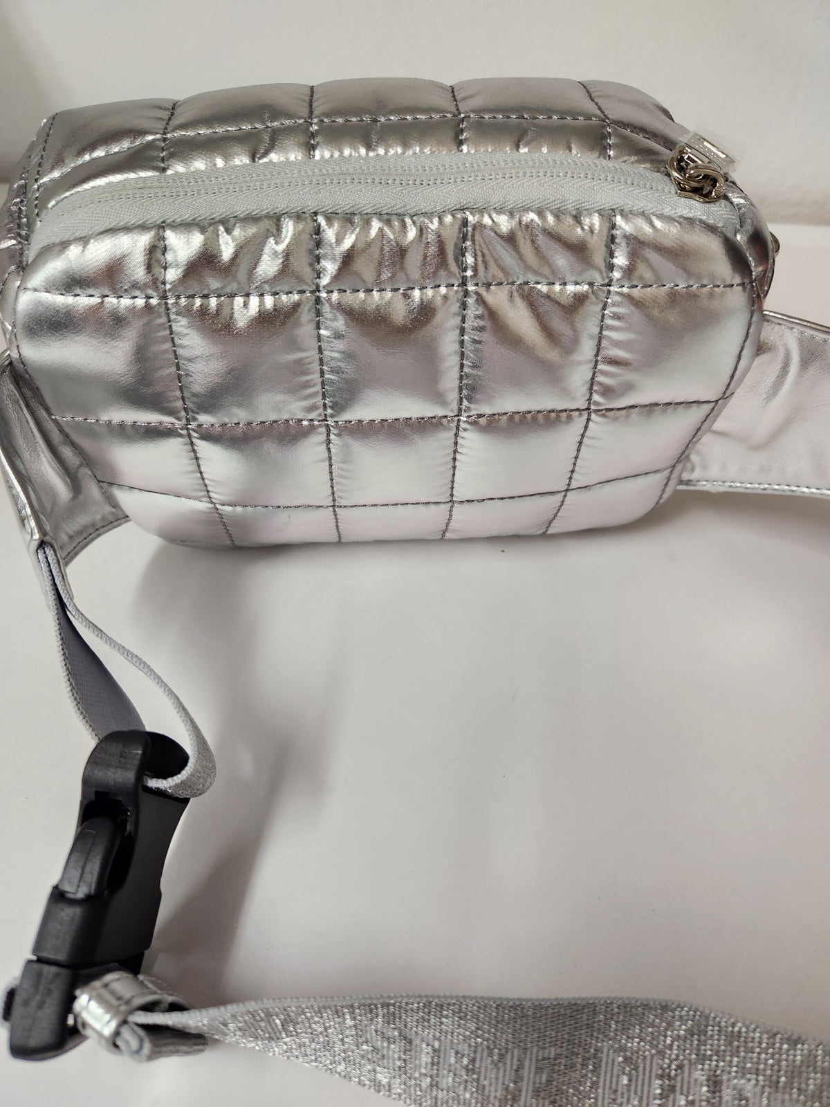 A metallic silver quilted crossbody bag with a zippered top and a matching fabric strap, shown on a plain light background.
