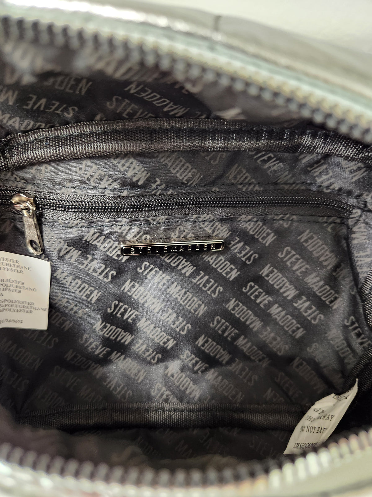 Close-up of the interior of a silver zippered bag, showing a black patterned lining with repeated 'Steve Madden' text and a small metallic logo plaque.