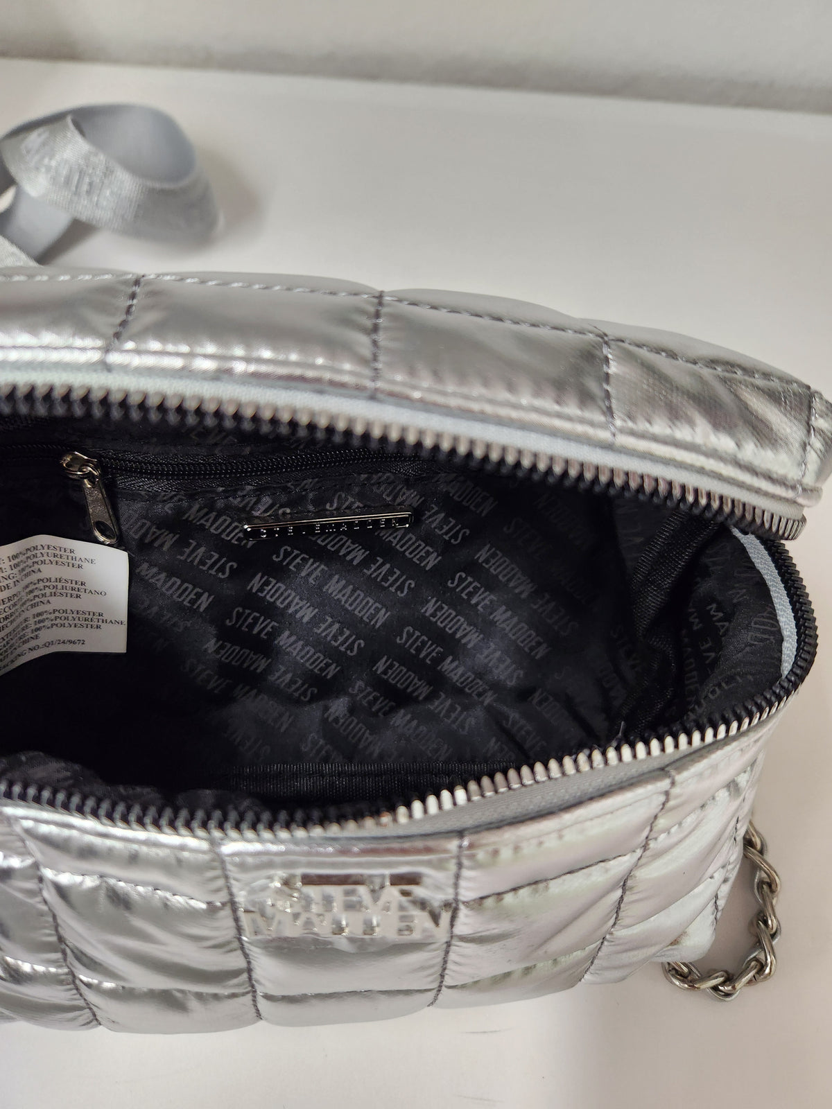 A close-up of a silver quilted metallic handbag shown open, displaying its black interior lined with the brand name 'STOCKMANN' in white lettering. A silver chain strap is visible on the right side.