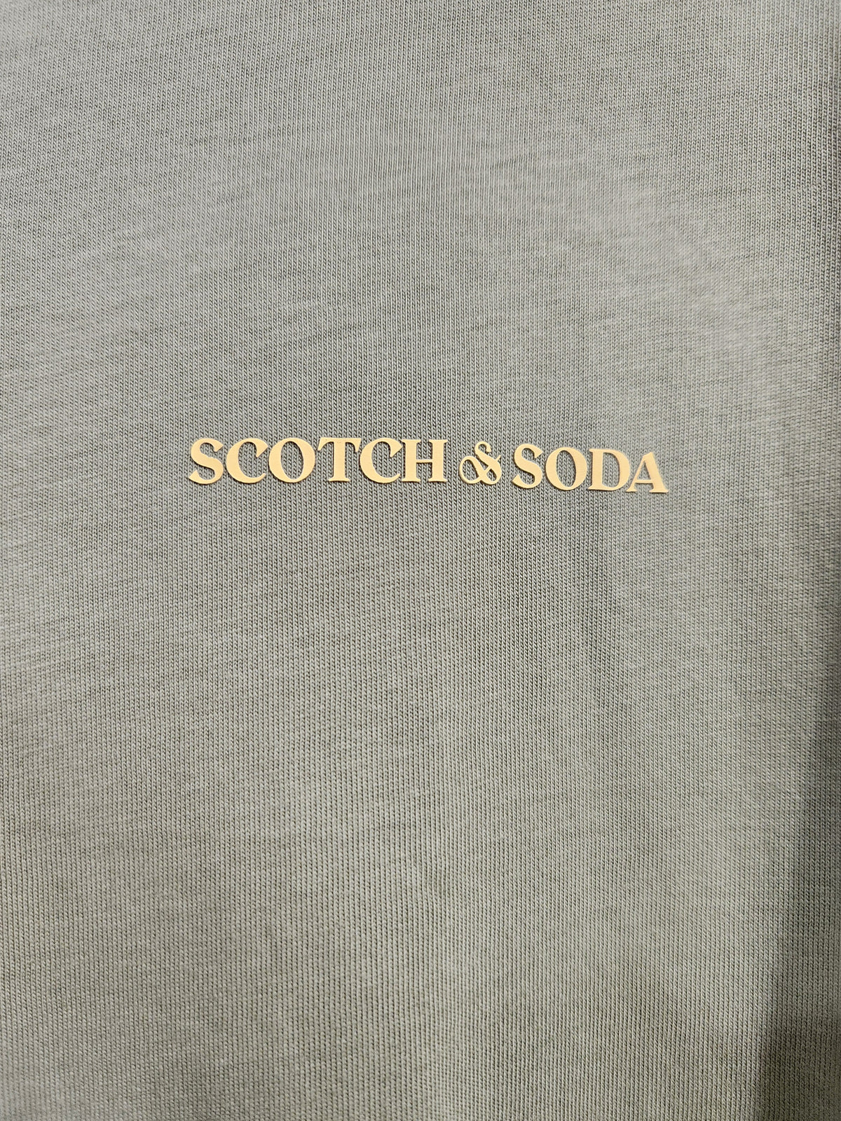 Close-up of a gray knit fabric with the words 'SCOTCH & SODA' printed in a light beige or cream tone across the center.