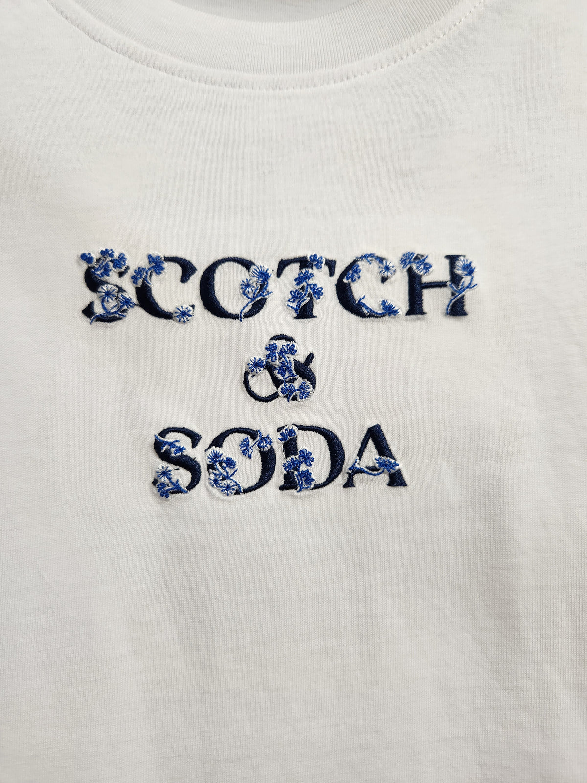 Close-up of a white crewneck t-shirt featuring the text 'SCOTCH & SODA' embroidered in dark navy blue with a blue and white floral pattern.