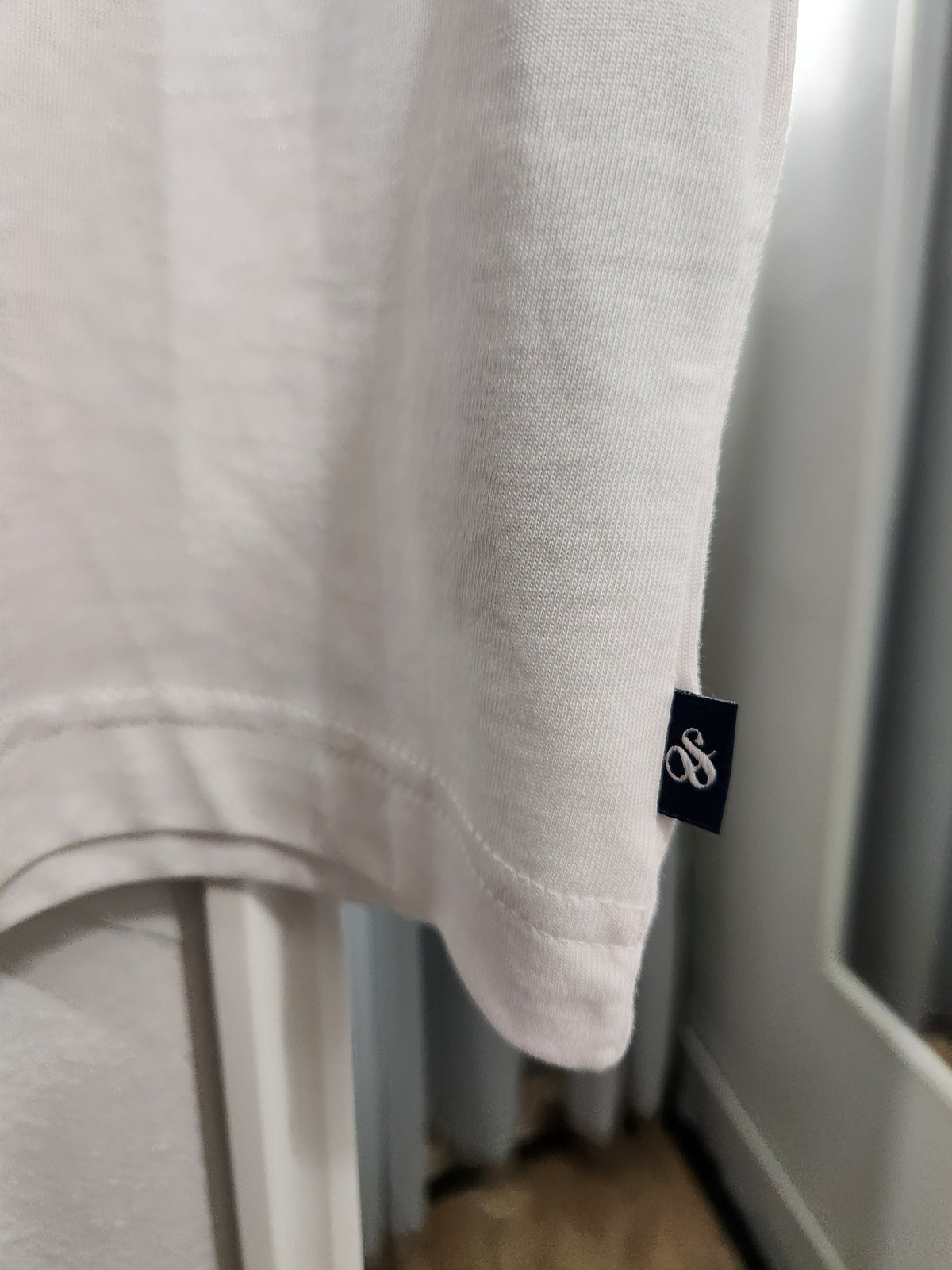 Close-up of a white cotton t-shirt sleeve showing a small black brand tag with a white logo, hanging next to a white chair.