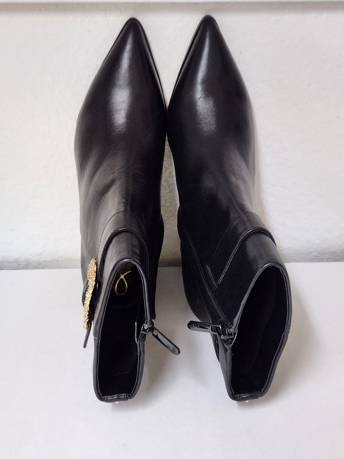 A pair of black leather pointed-toe ankle boots with side zippers and a gold-tone decorative accent on one boot, shown against a plain off-white background.