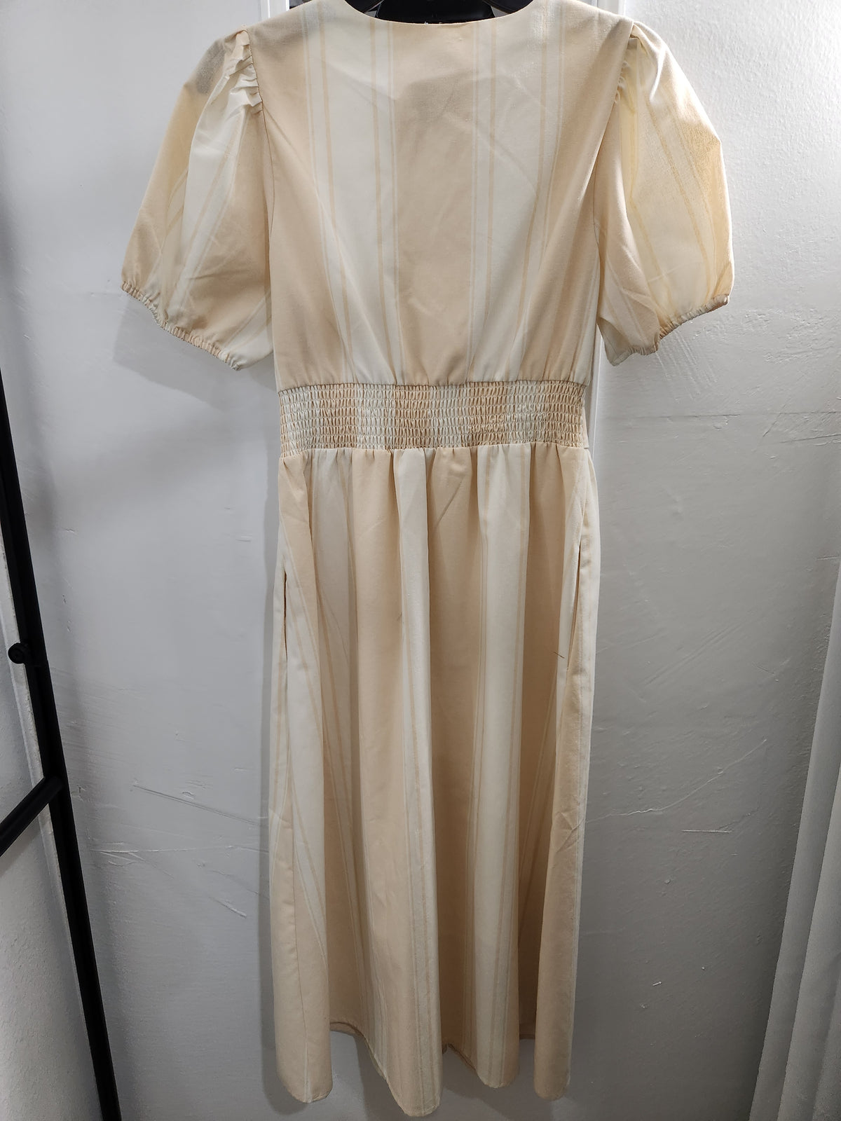A cream-colored midi dress with short puff sleeves and a smocked waistband, shown hanging against a light gray wall.