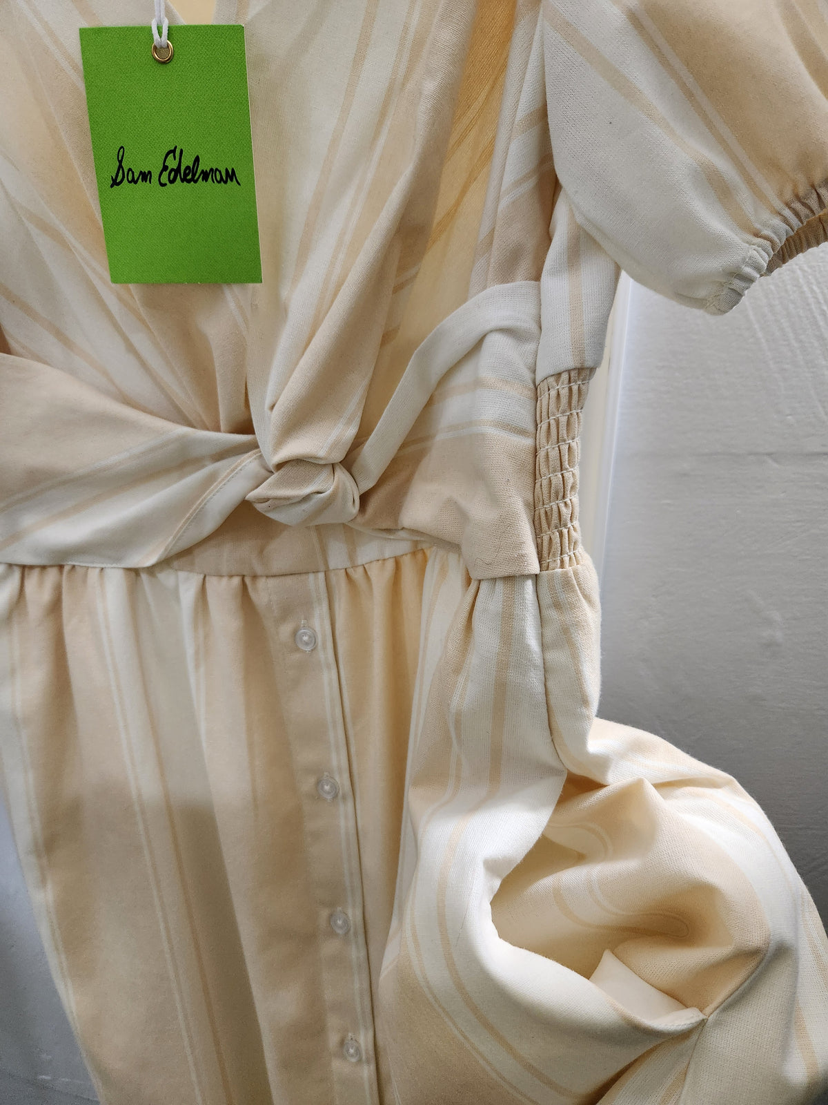 A close-up of a cream-colored button-front garment with gathered fabric and a knotted waist detail, shown against a light neutral background. A green hangtag with the name 'Jana Gobelman' is attached near the top.