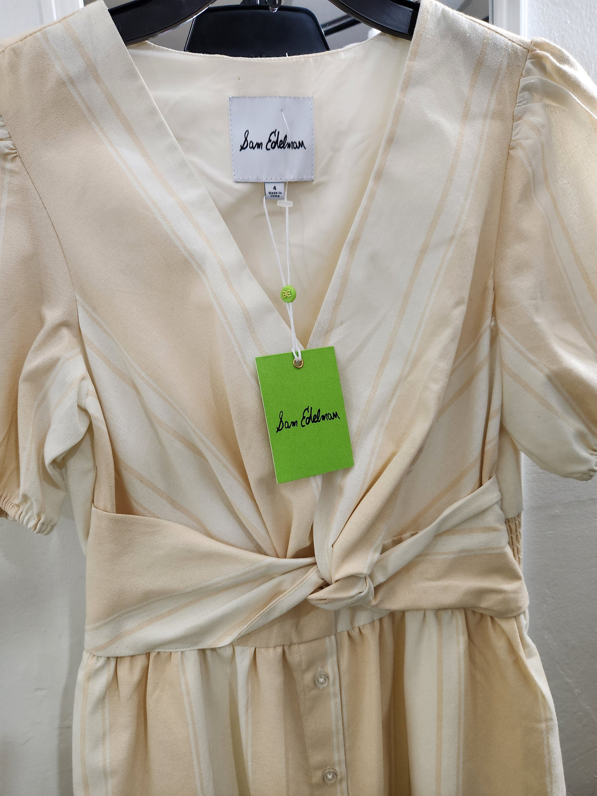 A cream-colored wrap dress with vertical stripes, featuring puff sleeves, a V-neckline, and a self-tie waist, shown hanging on a black hanger with a green brand tag.