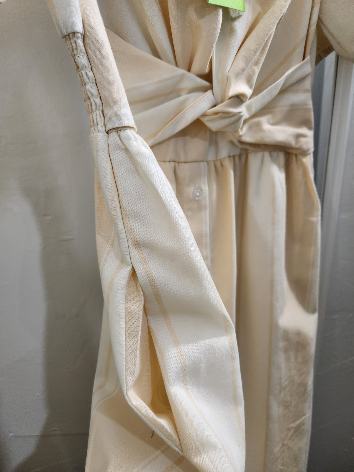 A close-up of a cream-colored wrap dress or tunic with a knotted waist and a small button detail, hanging against a neutral gray background.