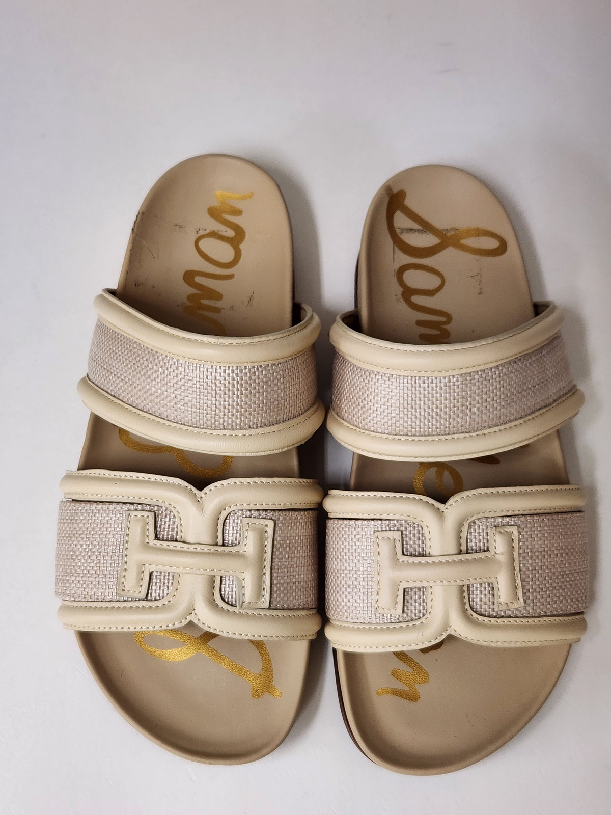 A pair of women's slide sandals with beige woven straps and a large beige H-shaped buckle detail, featuring a gold signature logo printed on the footbed.