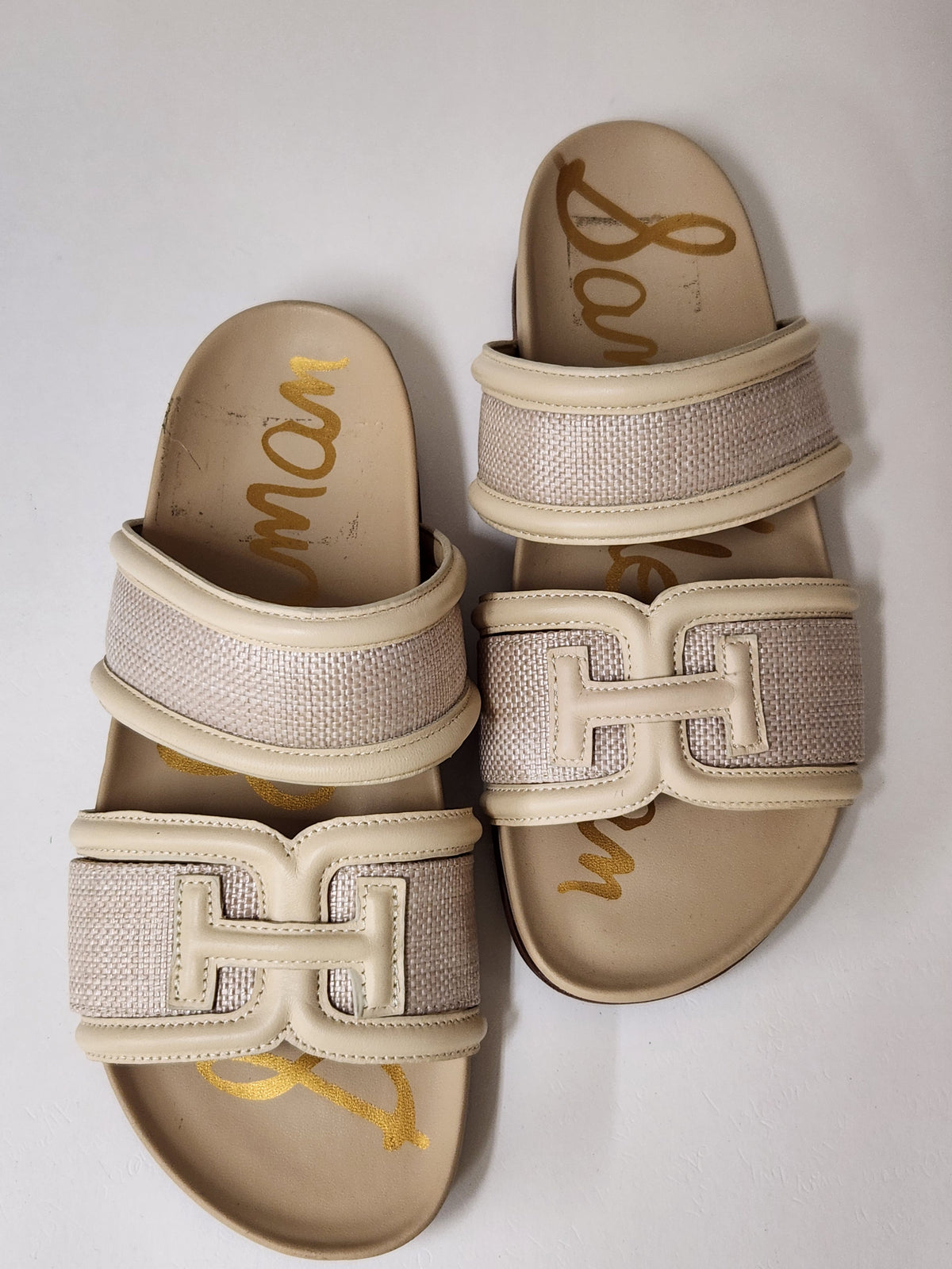 A pair of beige and cream slide sandals featuring a large interlocking H design on the straps, with textured woven panels and gold script branding printed on the footbeds.
