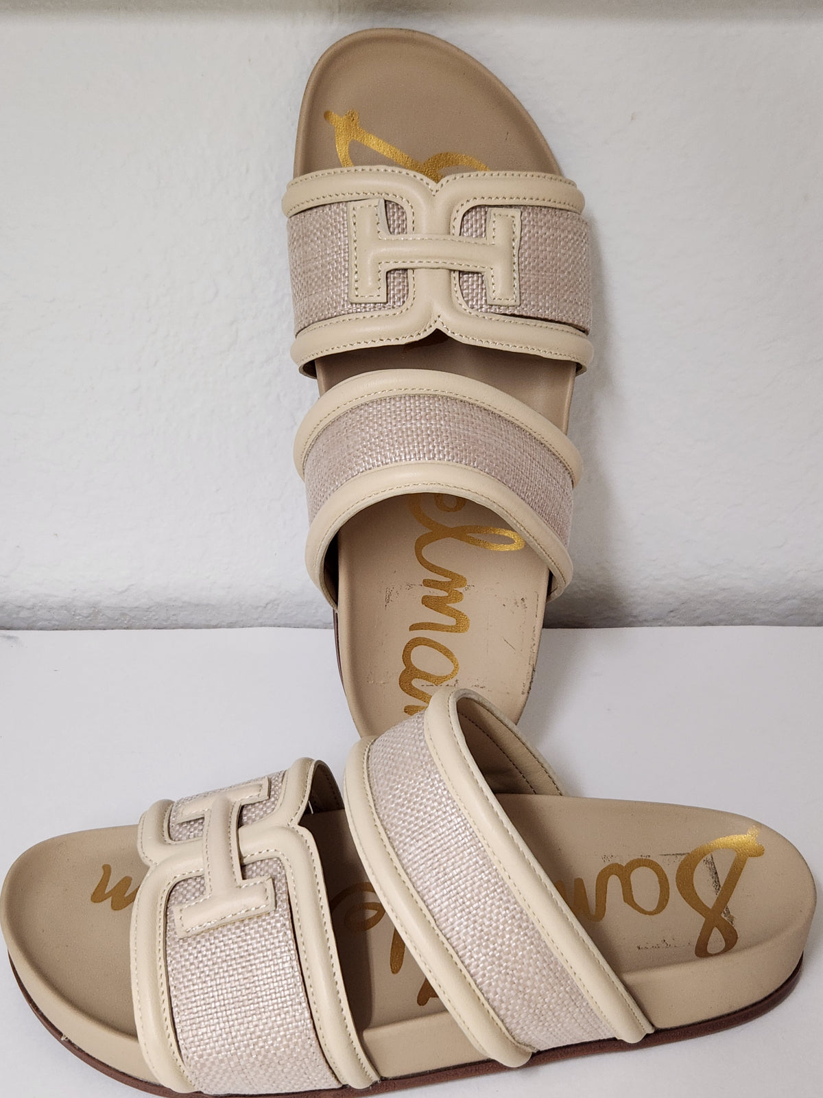 A pair of beige slide sandals with wide woven straps and a large decorative H-shaped buckle detail, displayed on a white surface against a white wall.