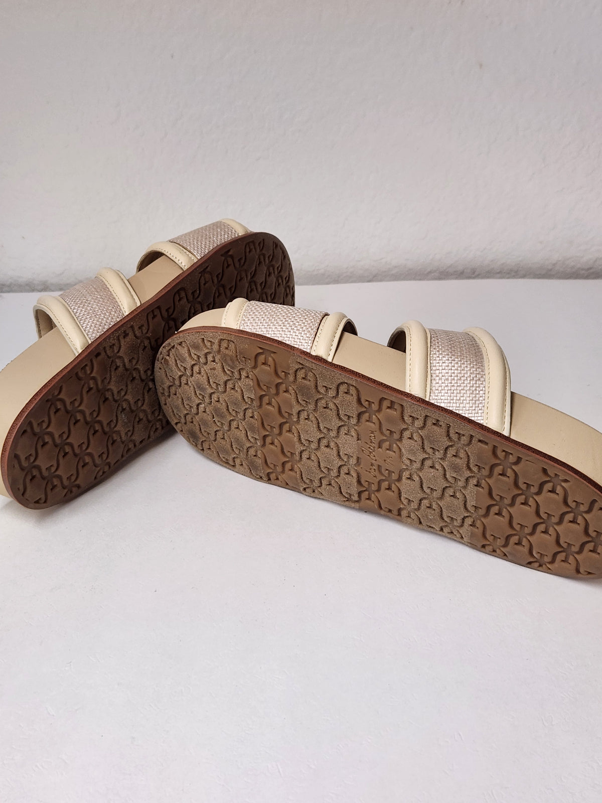 A pair of beige slide sandals with textured brown soles, displayed on a light gray surface against a white wall.