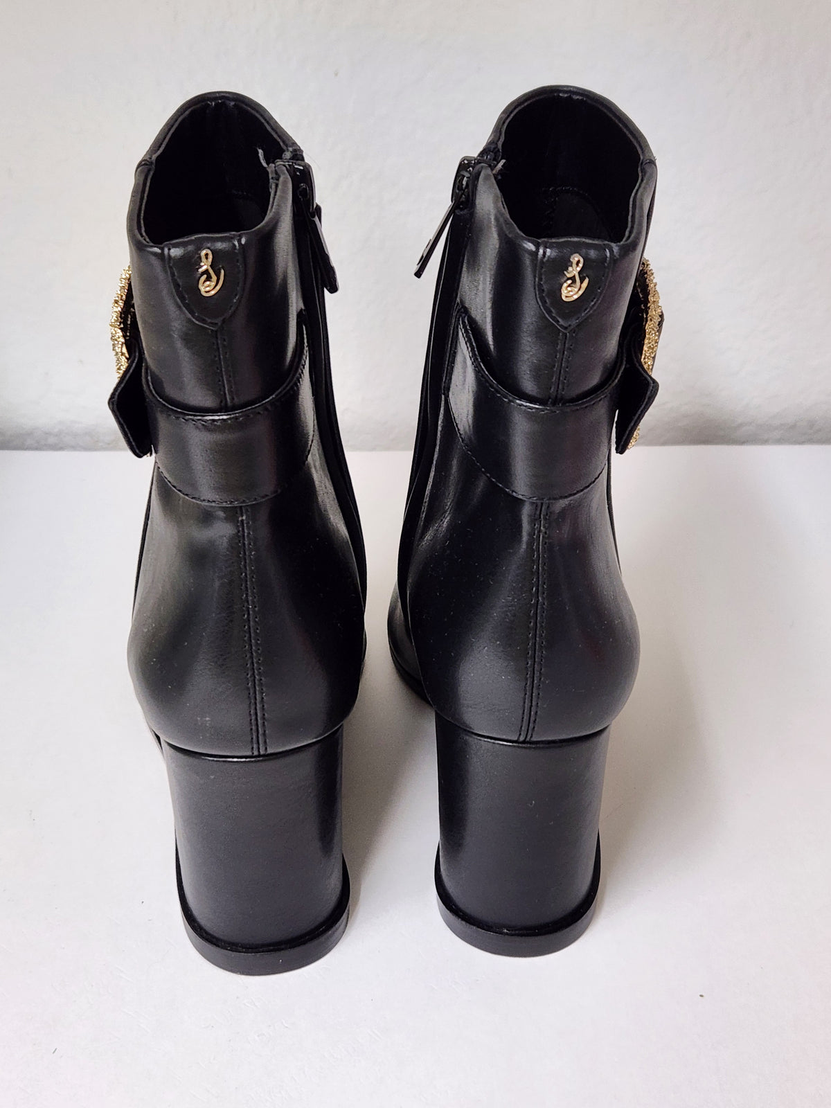 A pair of black leather ankle boots with block heels and gold-tone decorative hardware, shown from the back on a white surface against a white wall.