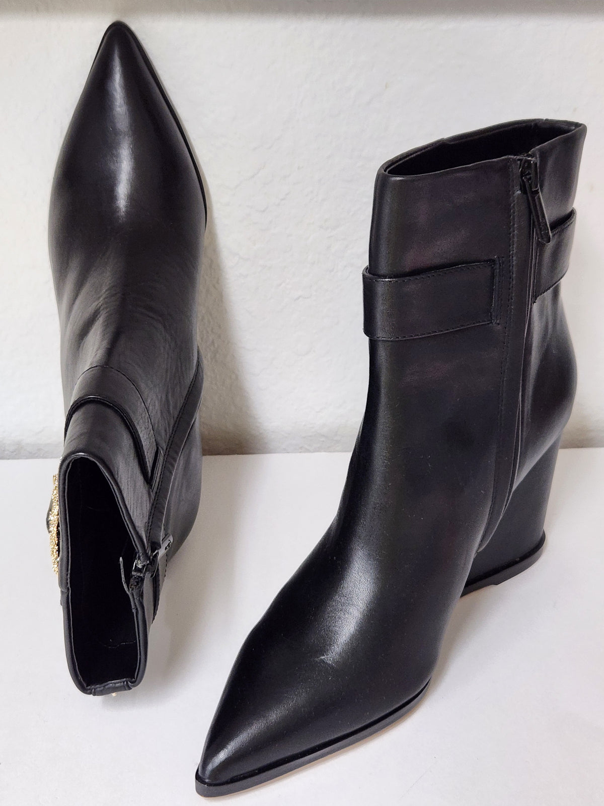 A pair of black leather pointed-toe ankle boots with a side buckle strap and a gold-tone rhinestone-embellished square buckle, displayed on a light gray surface.