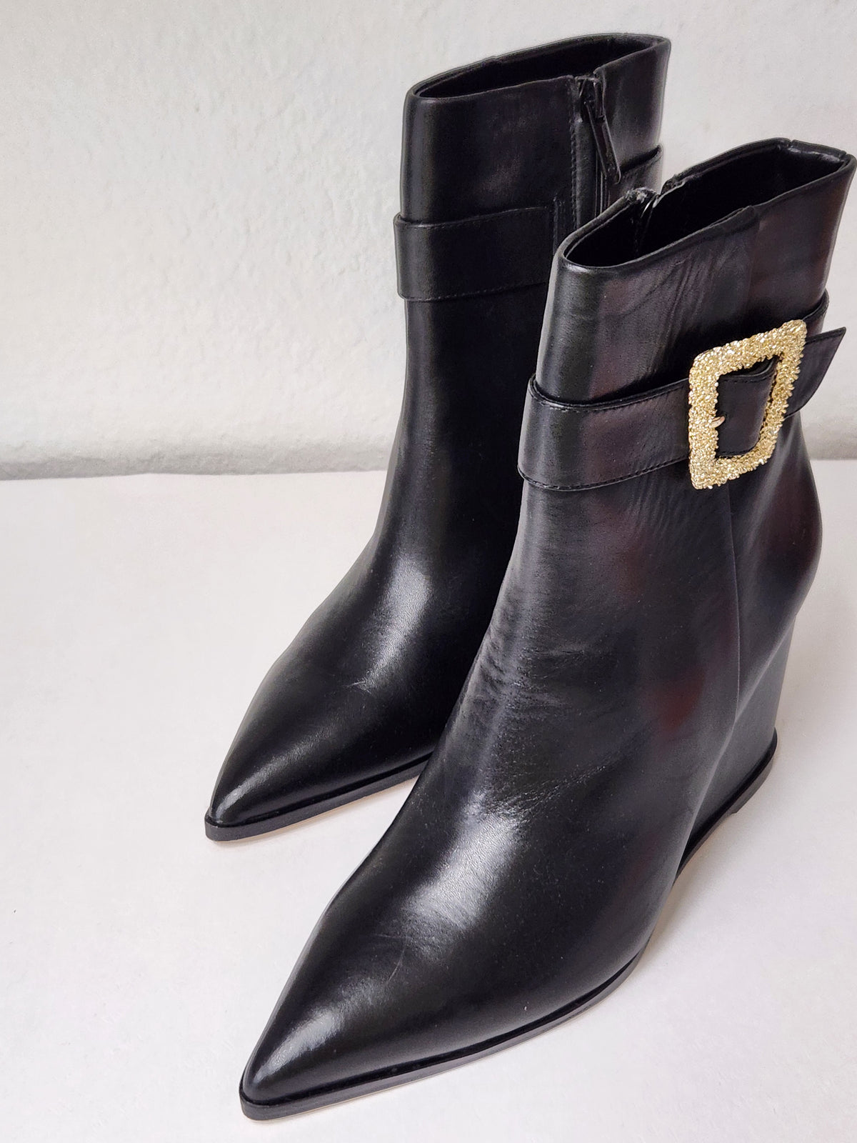 A pair of black leather pointed-toe ankle boots with a side buckle strap and a gold-tone rhinestone-embellished square buckle, displayed on a light gray surface.