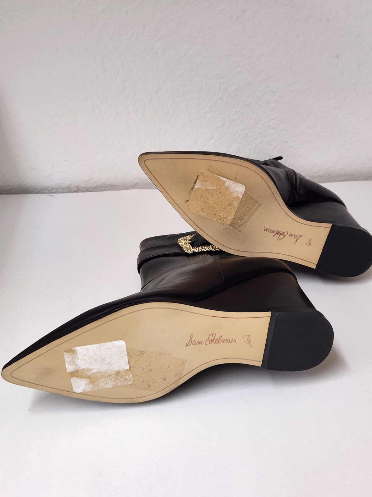 A pair of black leather pointed-toe ankle boots with a side buckle strap and a gold-tone rhinestone-embellished square buckle, displayed on a light gray surface.