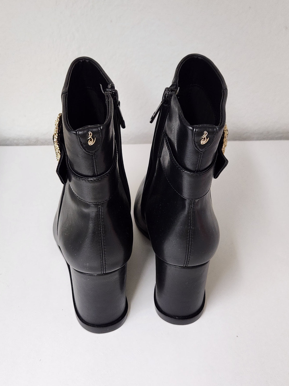 A pair of black leather ankle boots with block heels and gold-tone decorative hardware, shown from the back on a white surface against a light gray wall.