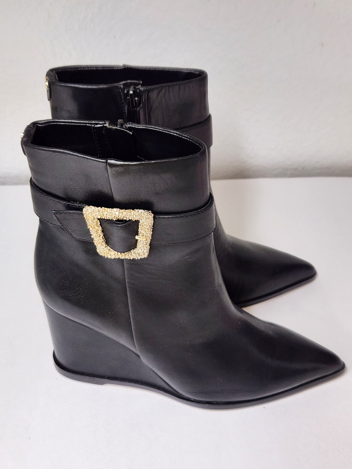 A pair of black leather ankle boots with pointed toes and wedge heels, featuring a decorative gold-tone buckle embellished with small clear rhinestones on the side, set against a plain light background.