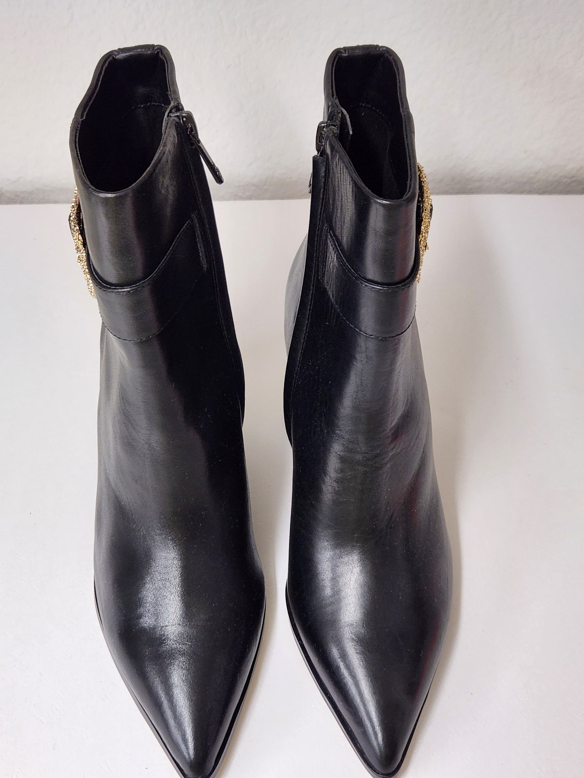 A pair of black leather pointed-toe ankle boots with side zippers and a gold-tone decorative embellishment near the collar, shown on a plain white surface.